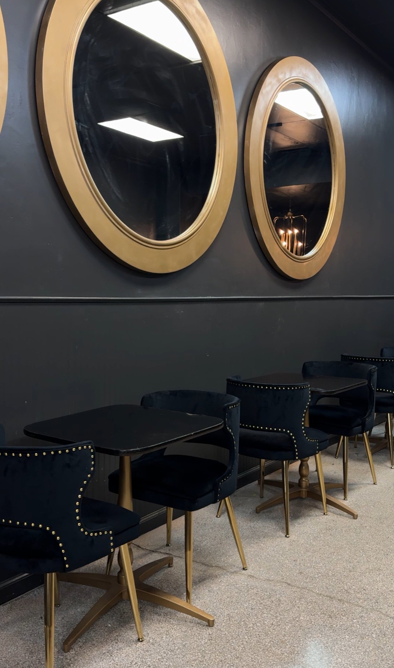 A room with black chairs featuring nailhead trim and gold legs, black tables, two round mirrors with gold frames hanging on a dark wall, and a light-colored carpet.