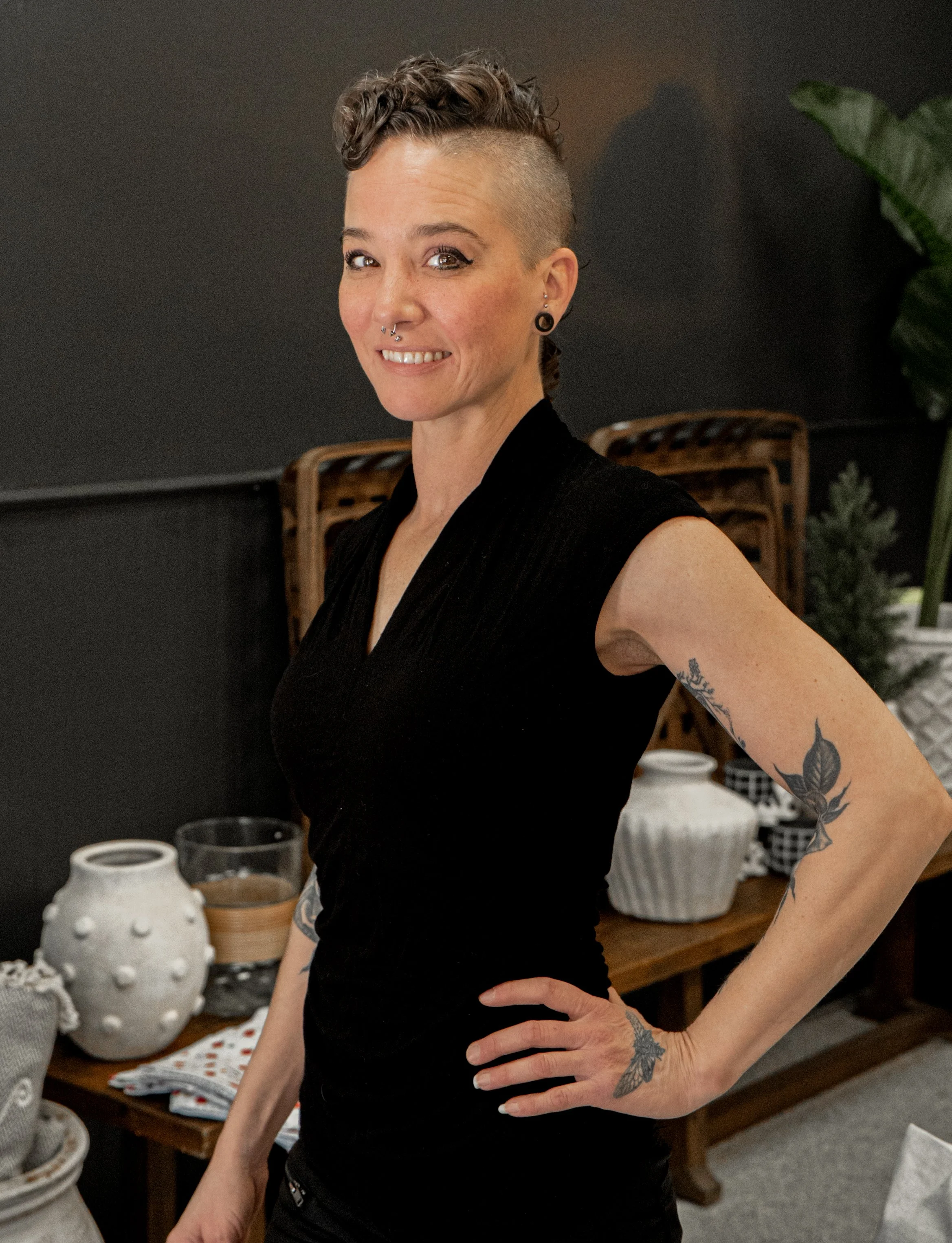 A woman with short curly hair and tattoos on her arm, wearing a sleeveless black top, smiling and standing in a room with pottery and plants in the background.