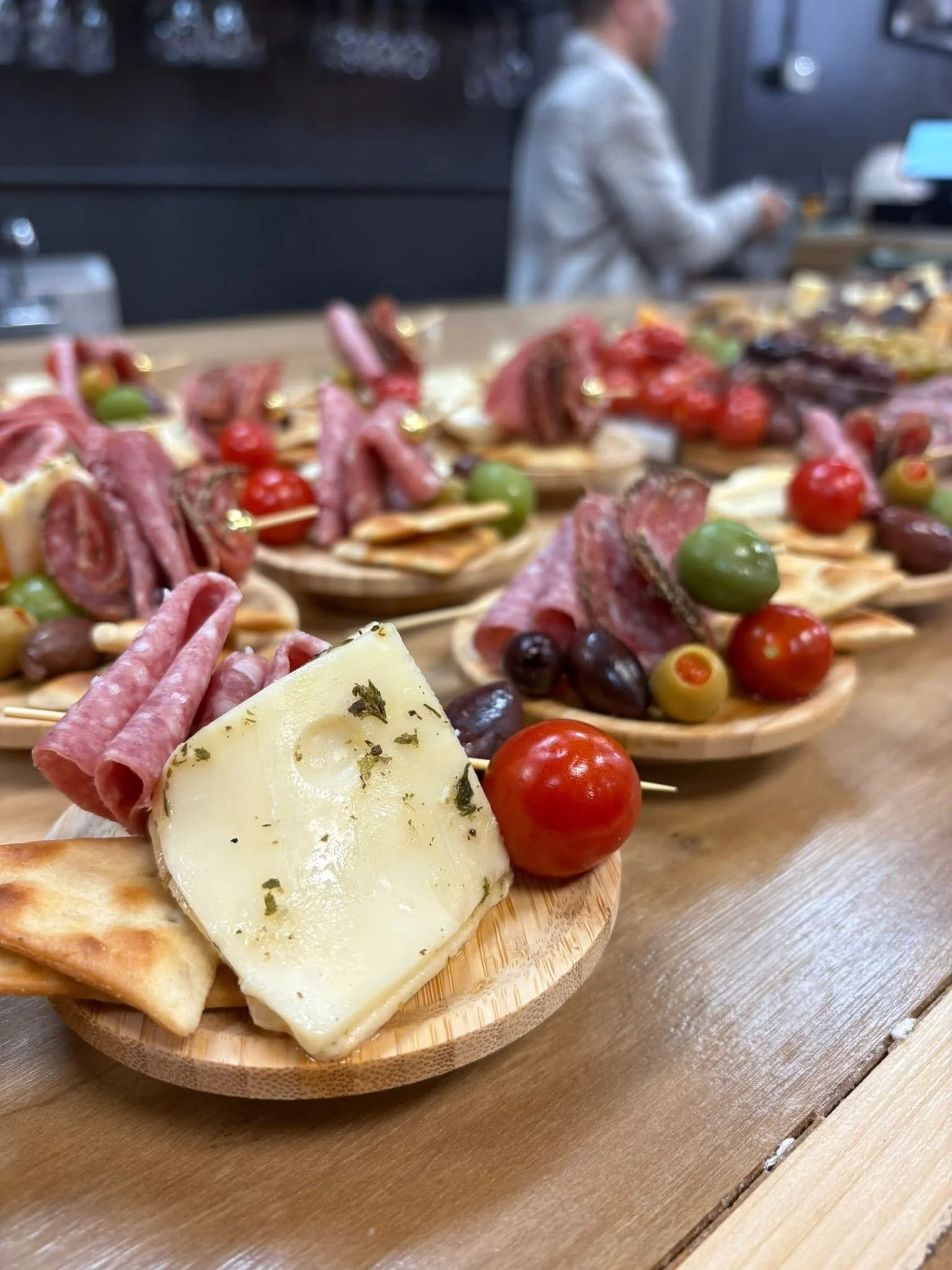 Assorted appetizers with cheese, cured meats, grapes, cherry tomatoes, and bread on wooden serving tray, with a blurred man in the background.
