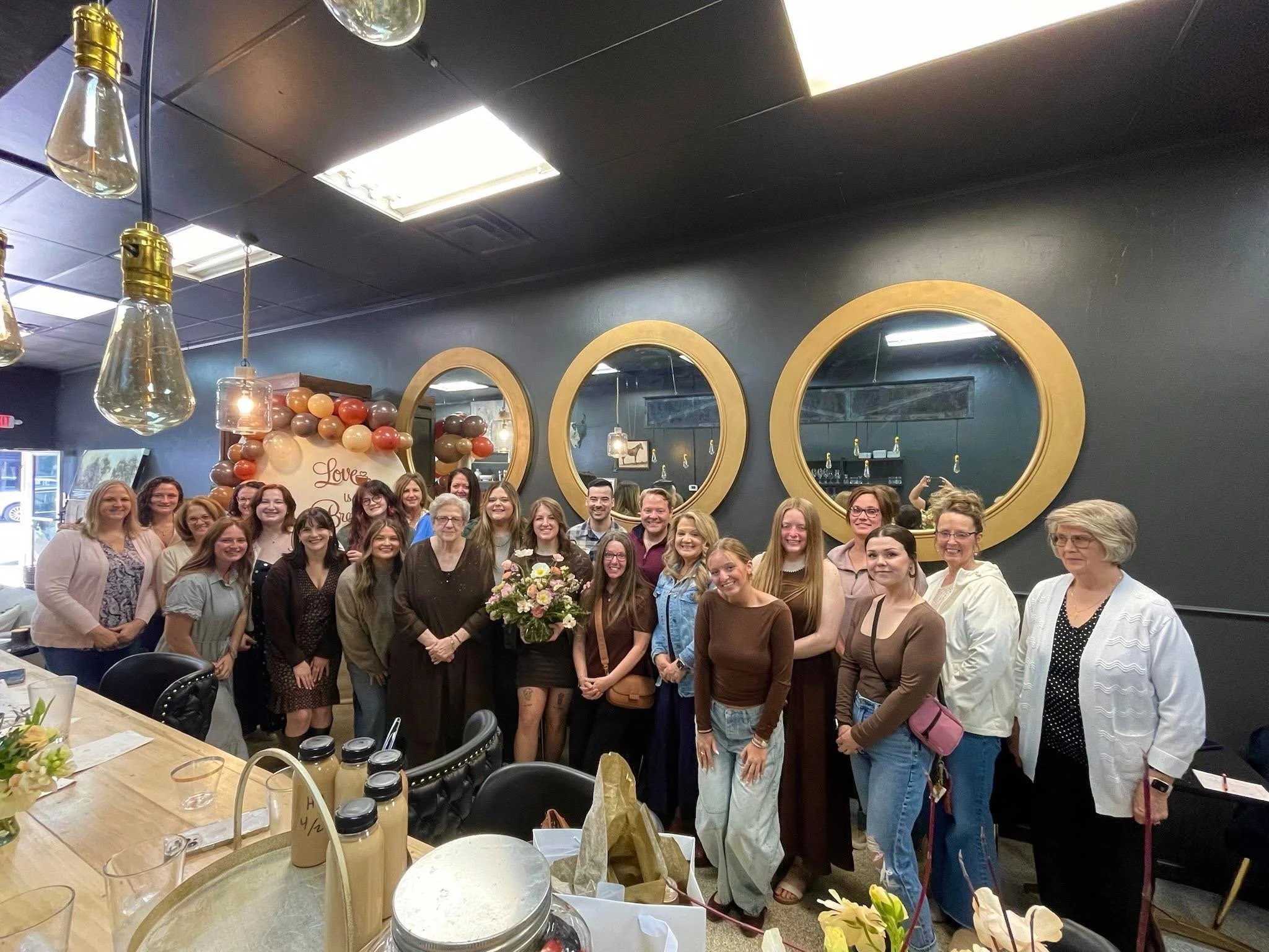 A large group of people gathered together in a restaurant or event space, smiling for a group photo. The room is decorated with balloons and mirrors on the wall, with a dark ceiling and bright lighting.
