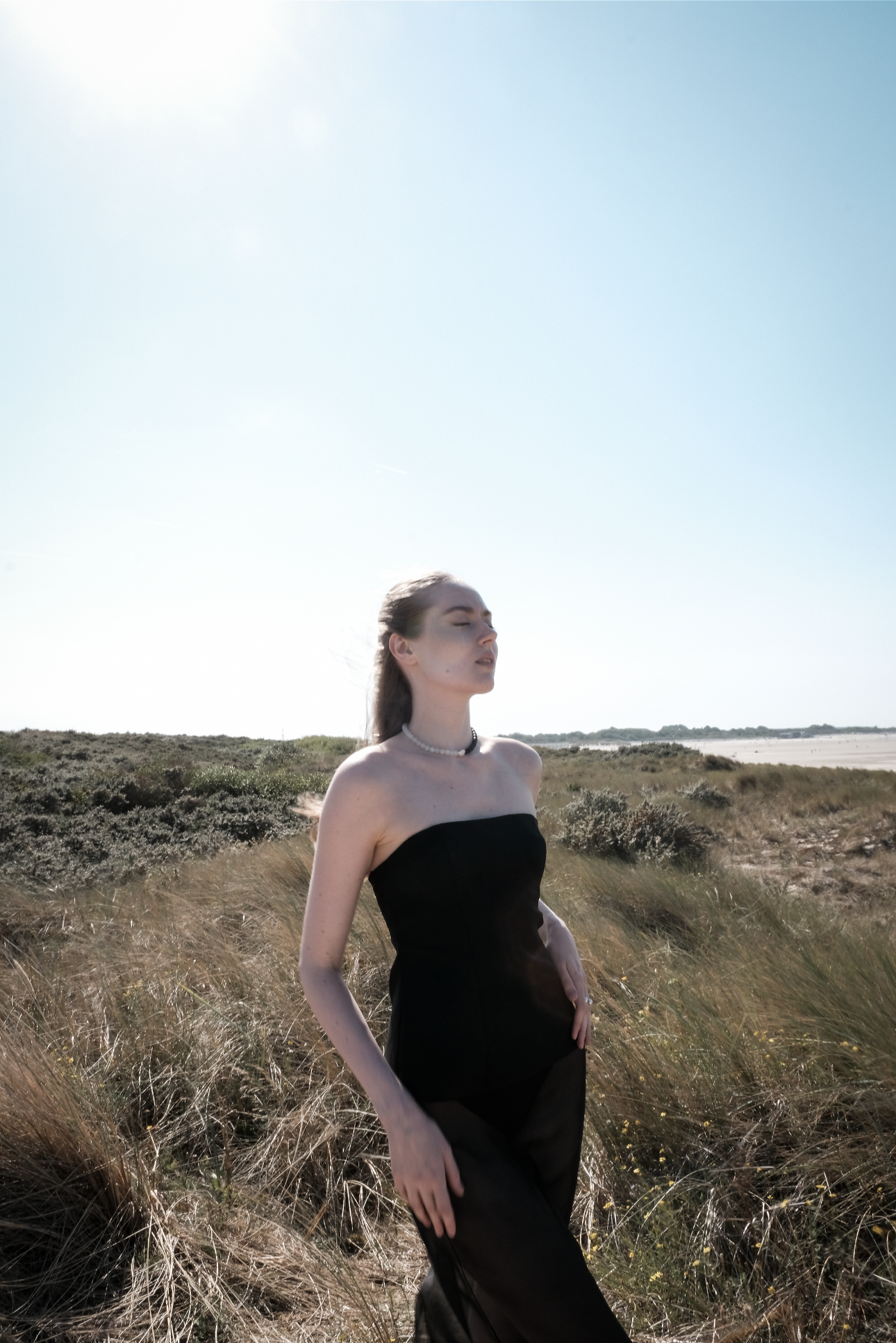 A young woman in a strapless black dress and pearl necklace standing in a grassy field under a clear sky