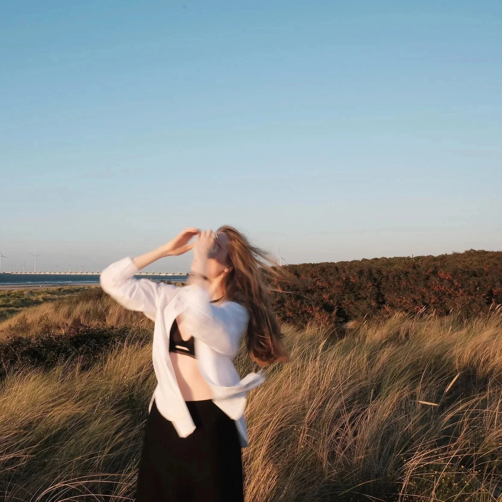 A woman standing in a grassy field near a beach with wind turbines in the background, her hair blowing in the wind as she holds her head with both hands during sunset.