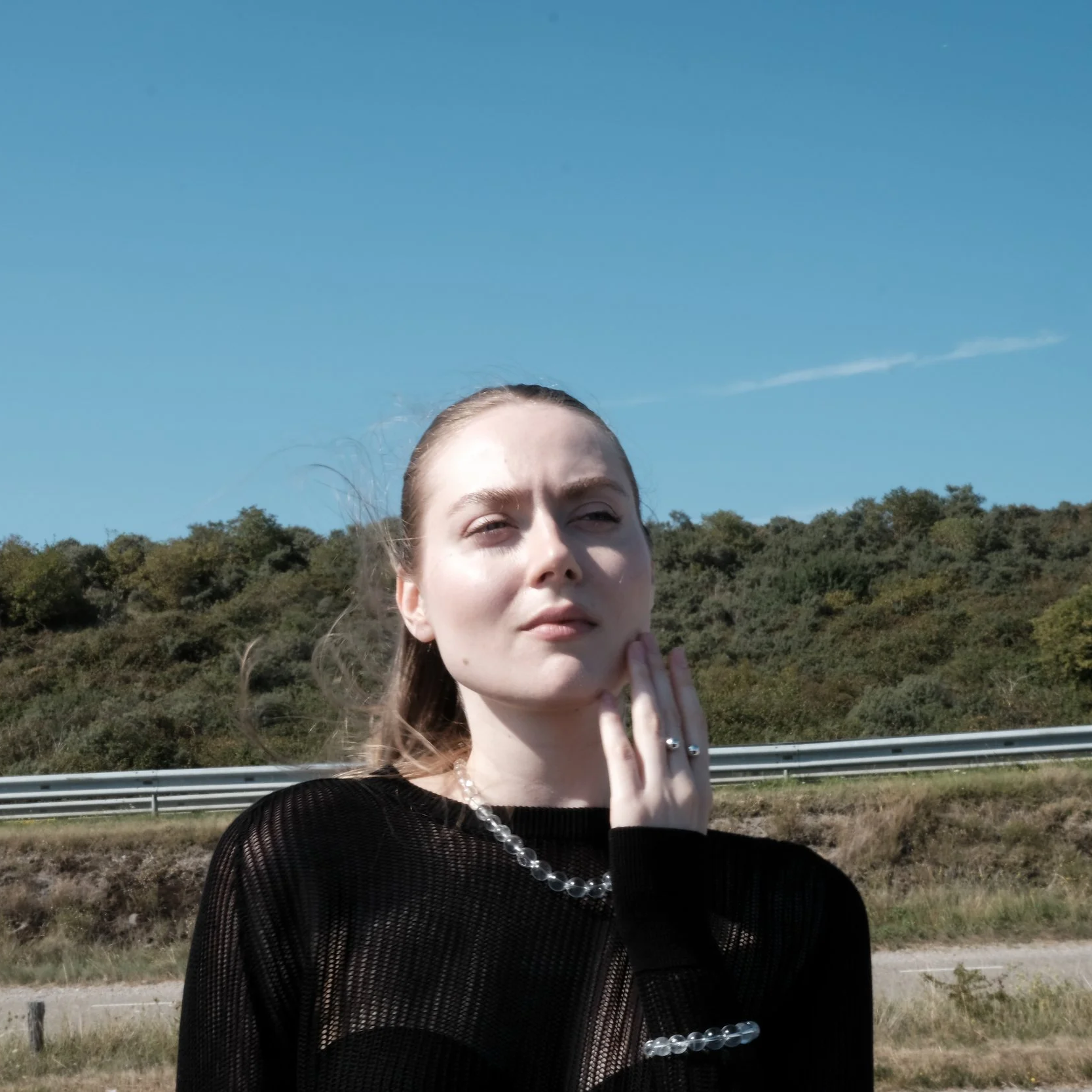 Young woman with long hair in a black mesh top, standing outdoors with a background of green hills, blue sky, and a highway guardrail, touching her chin with her left hand.