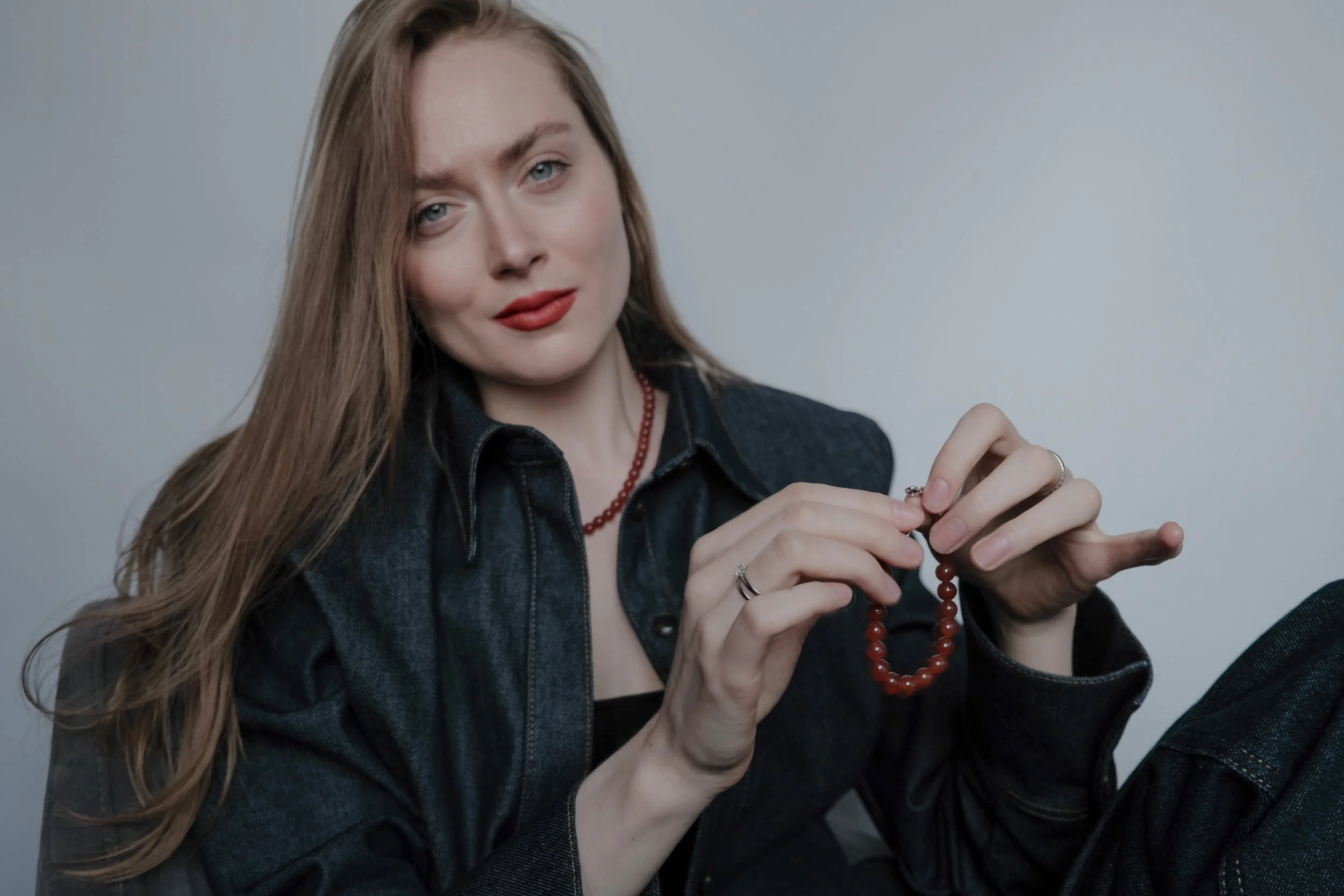 A young woman with long, light brown hair and blue eyes, wearing a black shirt, denim jacket, red lipstick, and red beaded jewelry, is sitting against a plain gray wall, holding a red beaded bracelet and looking at the camera.