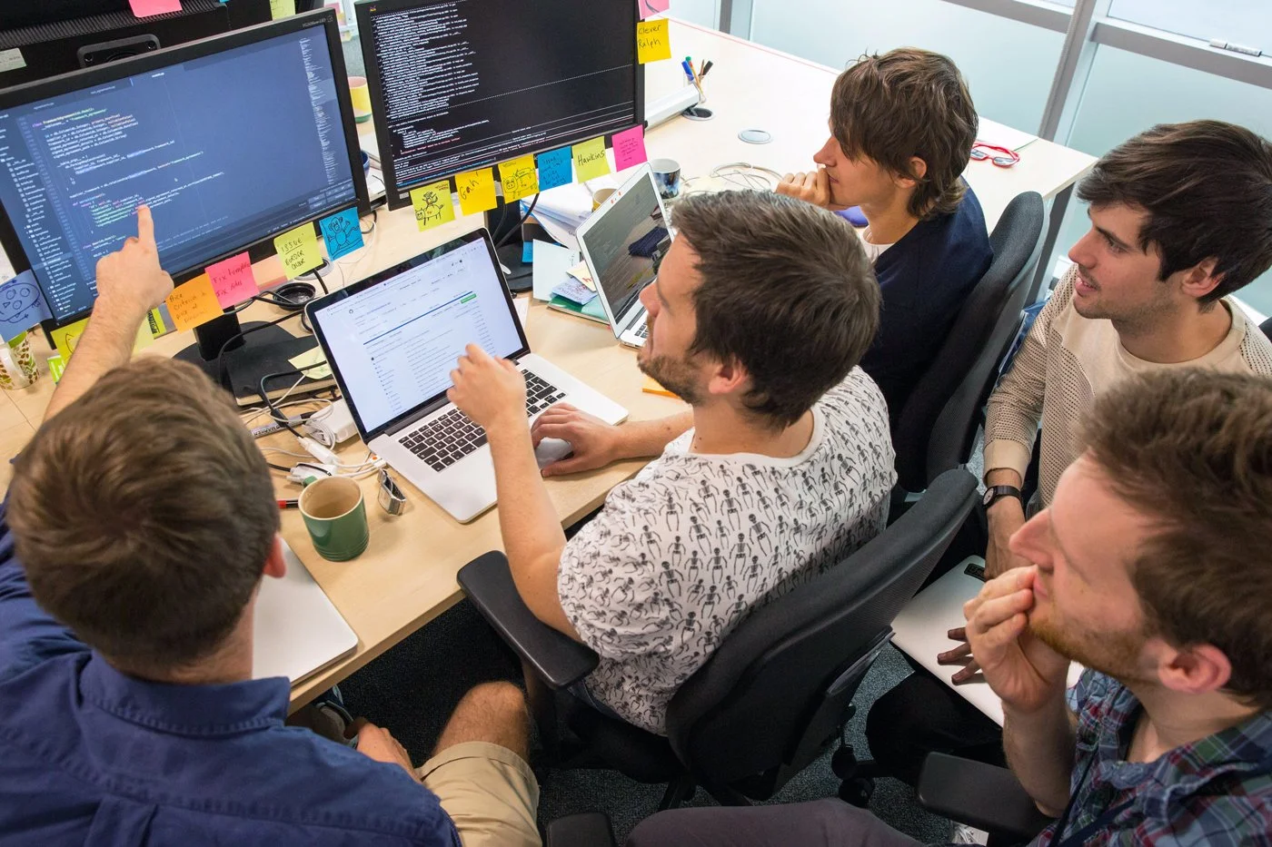 A group of six young men collaborating around a table in an office, with multiple monitors displaying code and a collaborative project, colorful sticky notes on the monitors and desk.