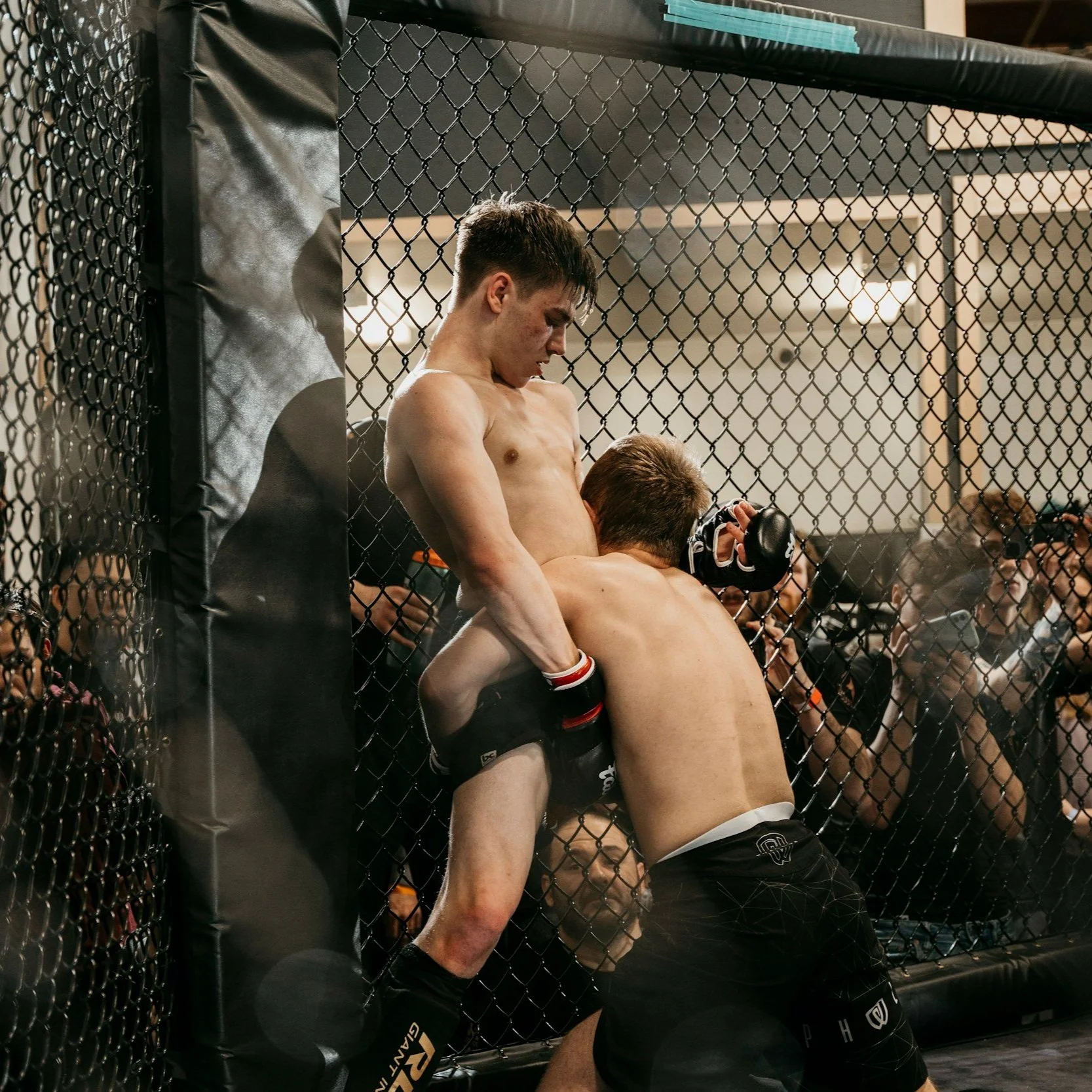 Mixed martial arts fighters inside a cage during a fight, with one fighter hitting the other against the chain-link fence, surrounded by spectators and photographers.