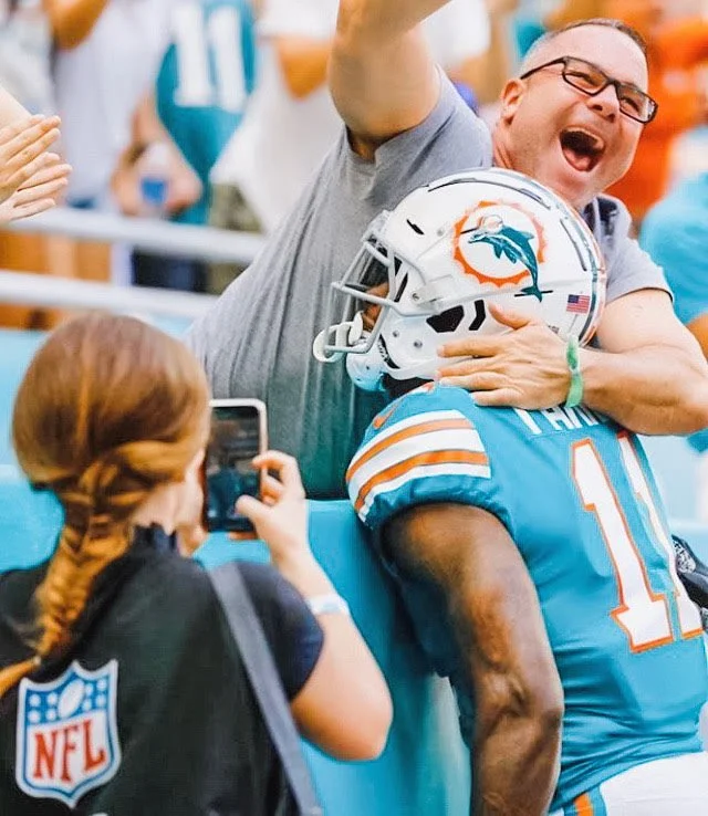 A woman with red hair recording a moment of celebration between a fan and a football player in Miami Dolphins gear, at a football game.