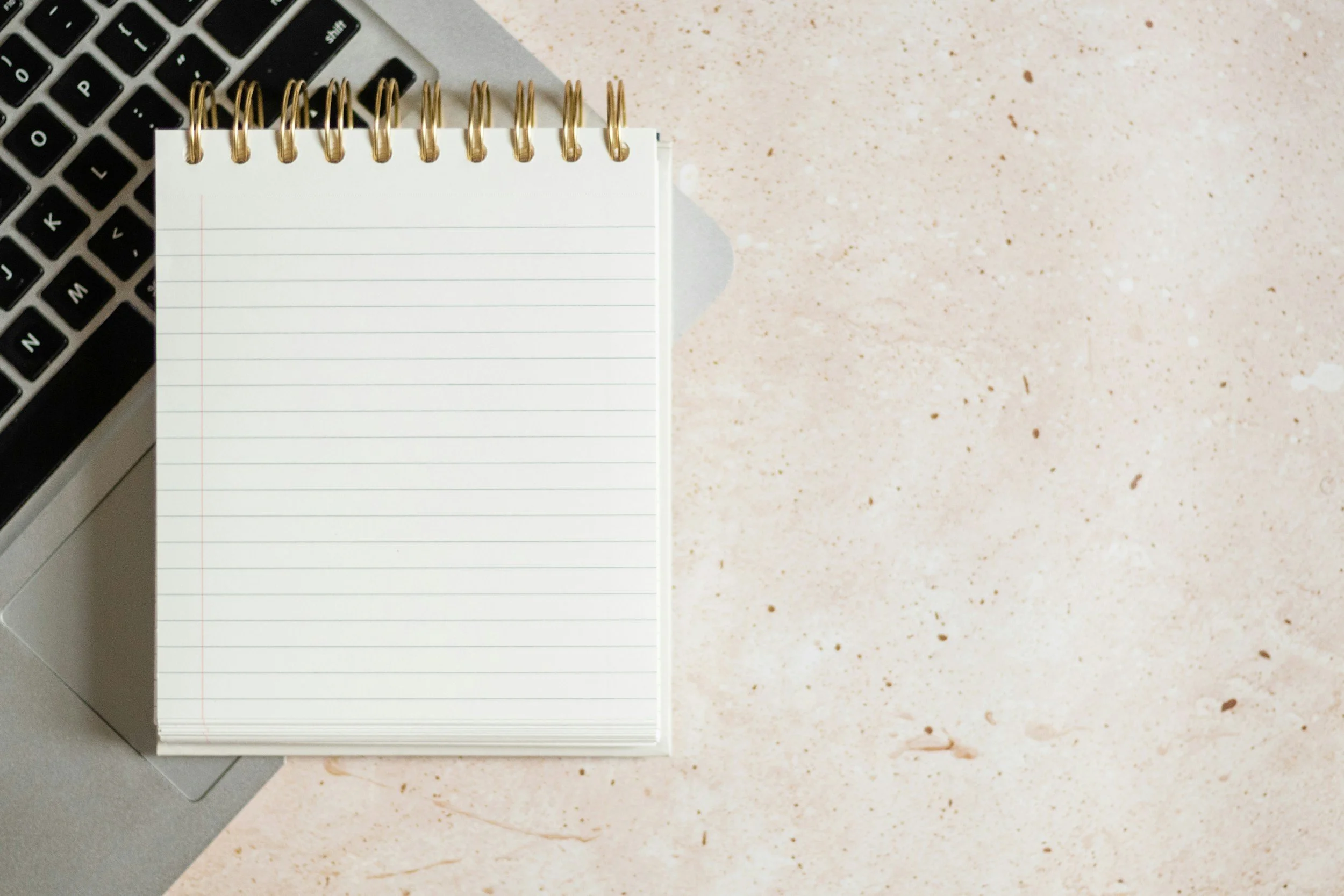A spiral notepad with blank lined pages next to a keyboard on a light-colored speckled surface.
