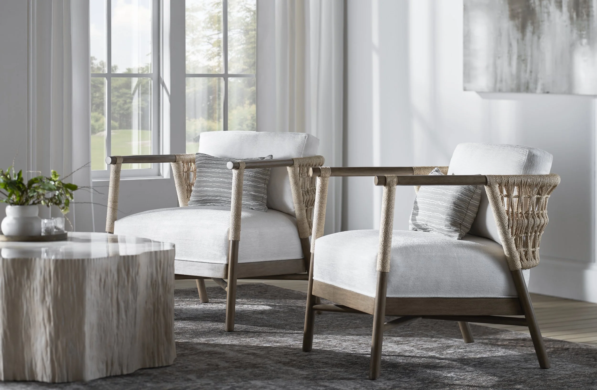 Living room with two light-colored armchairs, a wooden coffee table with a potted plant, large windows with white curtains, and abstract wall art.