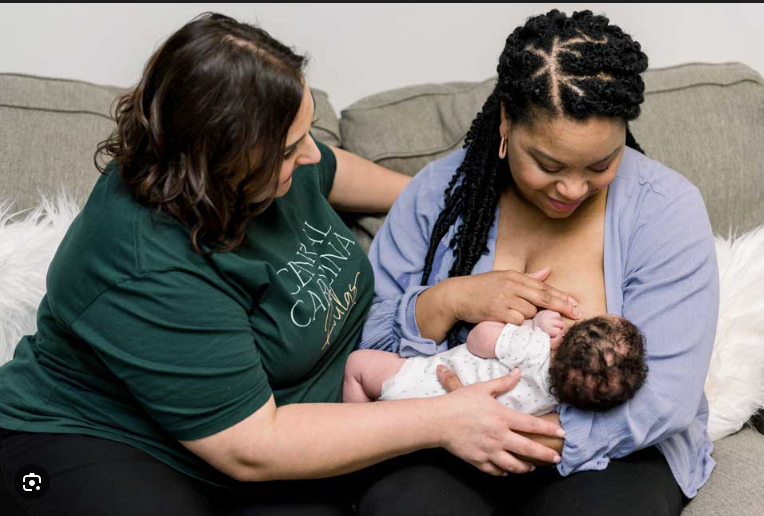 A woman with dreadlocks breastfeeds a newborn baby while sitting on a gray couch, with another woman in a green shirt supporting her.