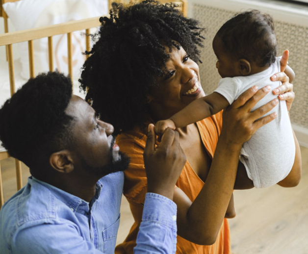 A woman holding a baby while a man playfully interacts with the baby in a cozy room.