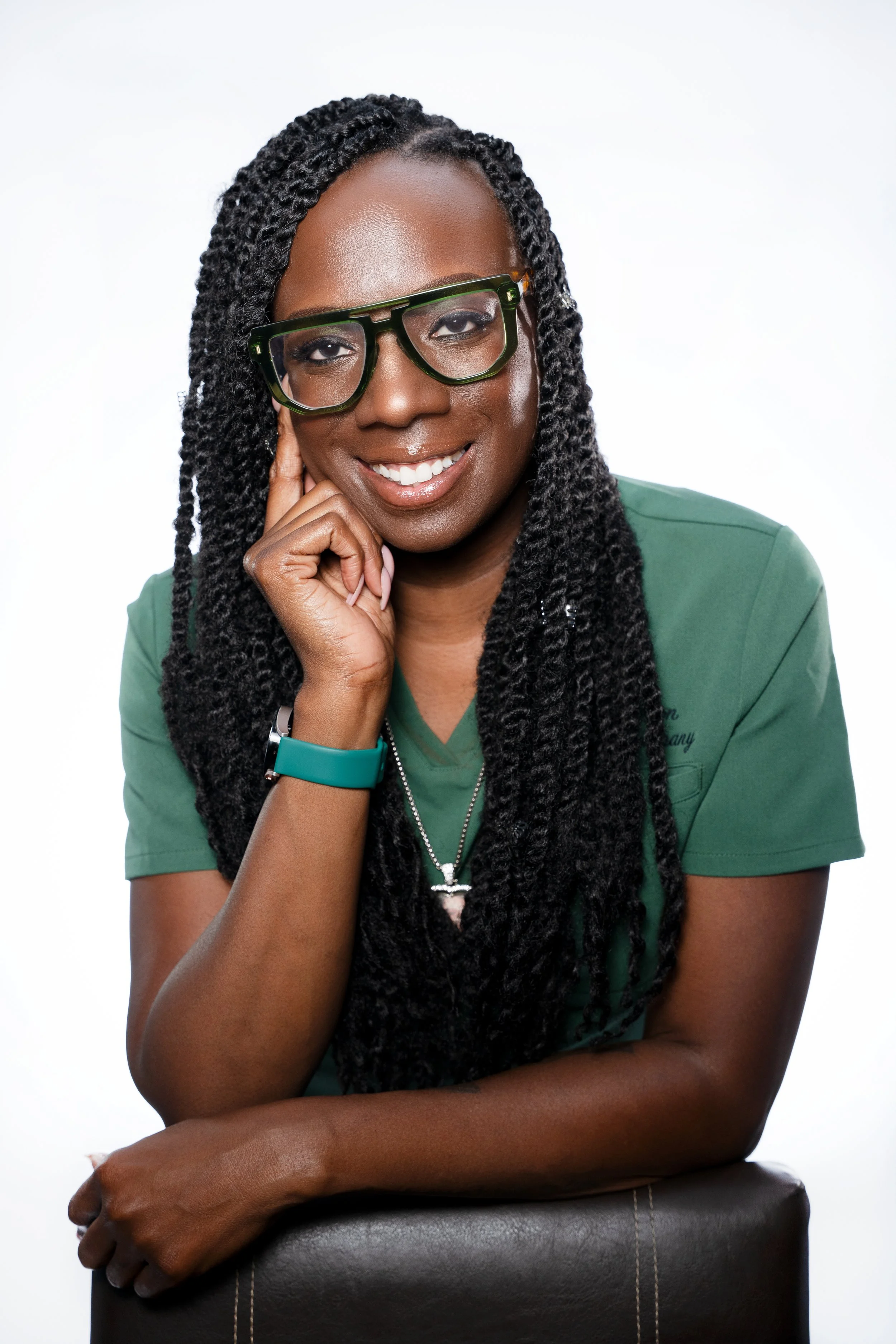 Portrait of a smiling woman with glasses, wearing a green shirt, resting her hand on her chin, against a white background.