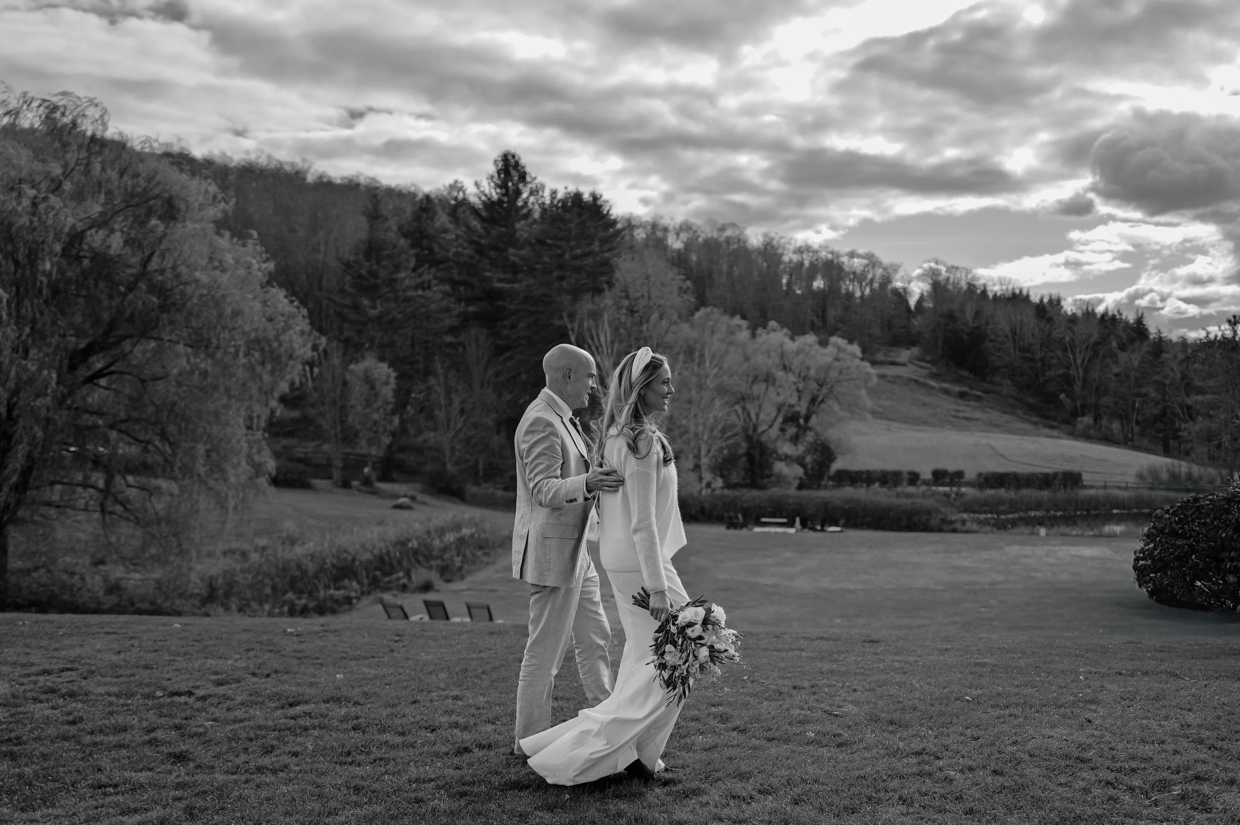 A black-and-white photograph of a bride and groom outdoors on a grassy field. The bride is holding a bouquet and wearing a white dress, while the groom, in a suit, stands beside her with his hand on her arm. Wedding photography and photographer