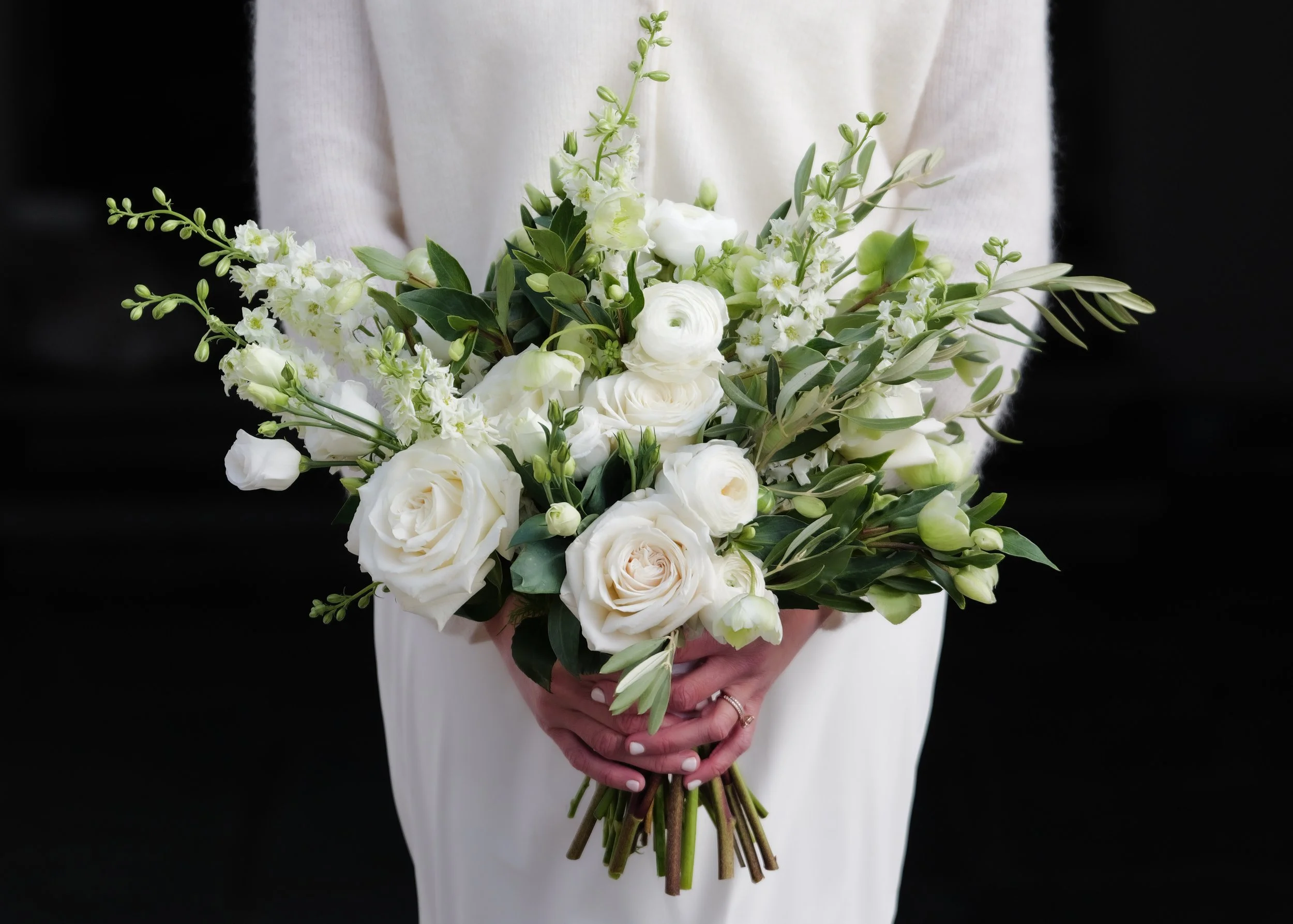 Person holding a bouquet of white roses, lisianthus, and greenery, dressed in white, against a black background.