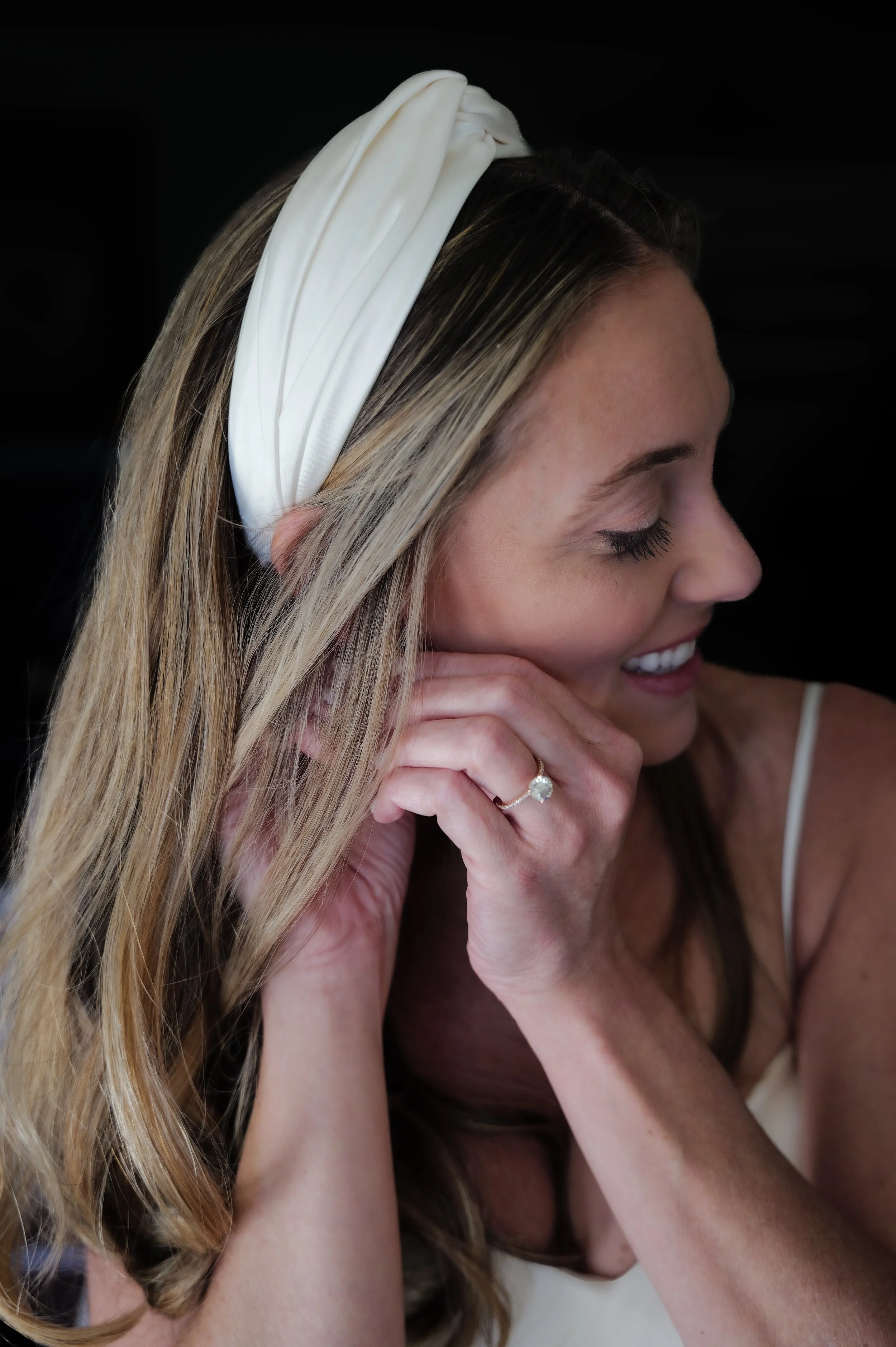 A bride with blonde hair and a white headband, smiling and looking down, touching her face with her right hand, showing a ring on her finger.