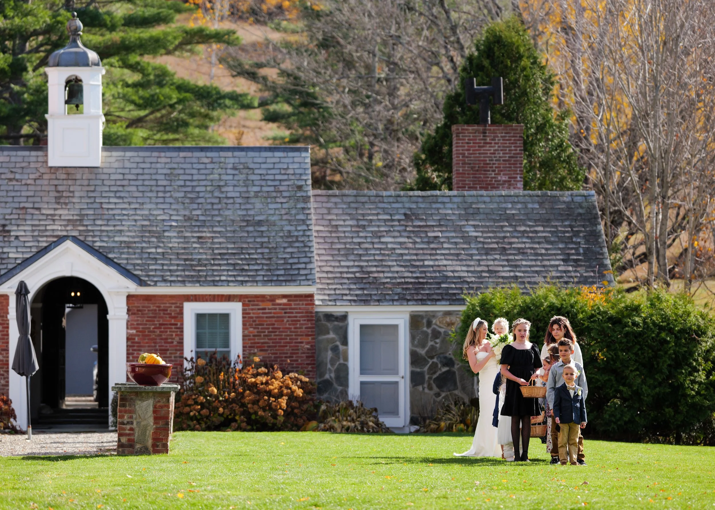 A group of six people, including a bride with a white dress, a woman with a headband, and four children, are gathered on a green lawn in front of a rustic house with brick and stone walls, a sloped roof, and a small bell tower on top, amid trees with