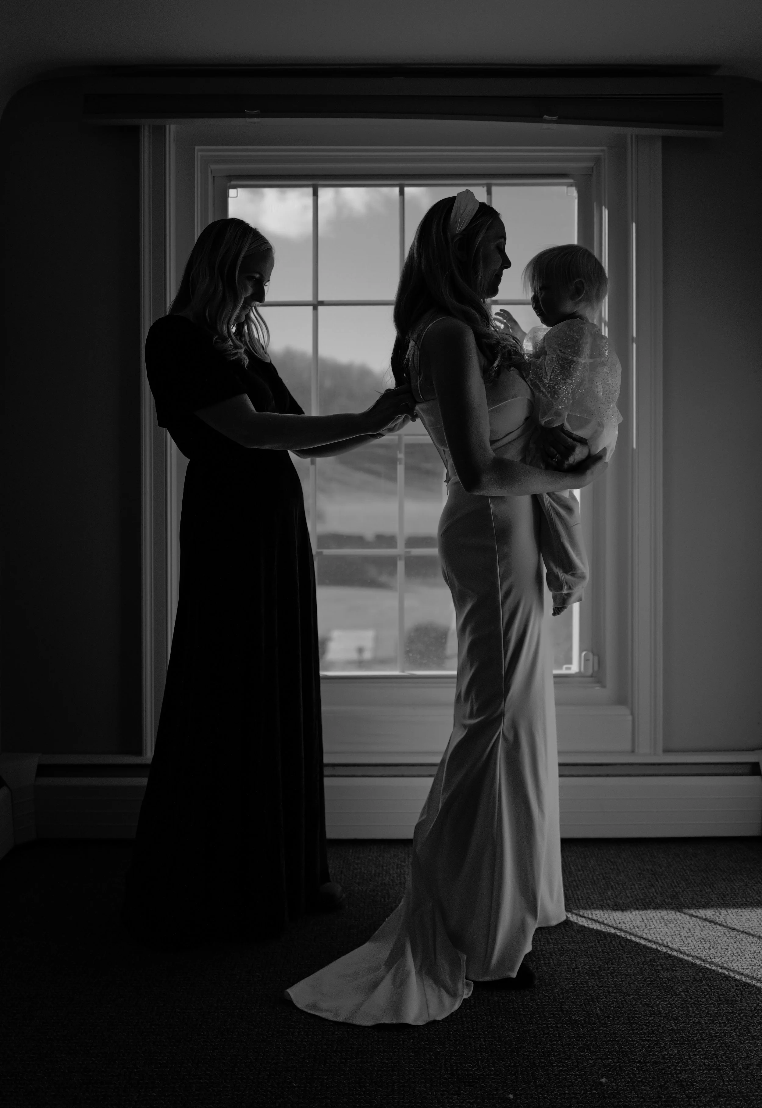 Silhouette of a woman in a wedding dress holding a young girl, with a woman adjusting the dress in front of a window.