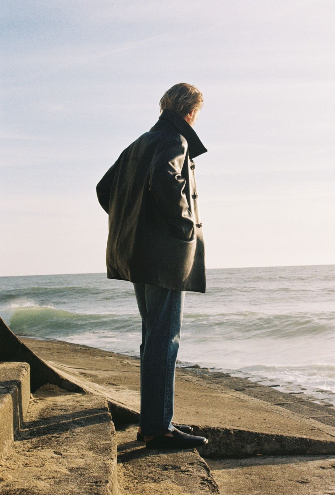 A person with hair tied back, wearing the Peace Uniform of Jeans, white T Shirt, black leather Deerskin Car Coat standing on a concrete seawall and looking out at the ocean during a cloudy day.