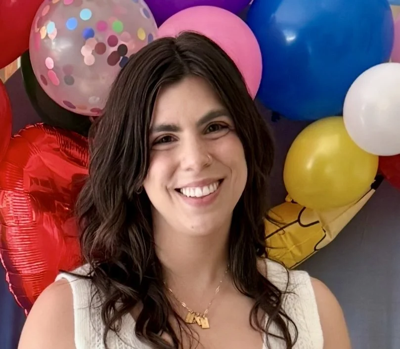 A woman smiling in front of colorful balloons, including pink, blue, yellow, white, and transparent balloons with confetti inside.