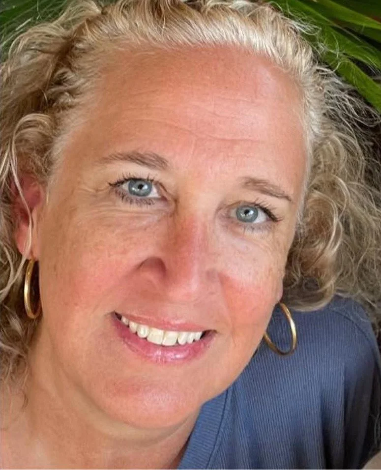 Close-up of a woman with curly blonde hair, blue eyes, wearing gold hoop earrings, smiling, and wearing a dark blue top, with green foliage in the background.