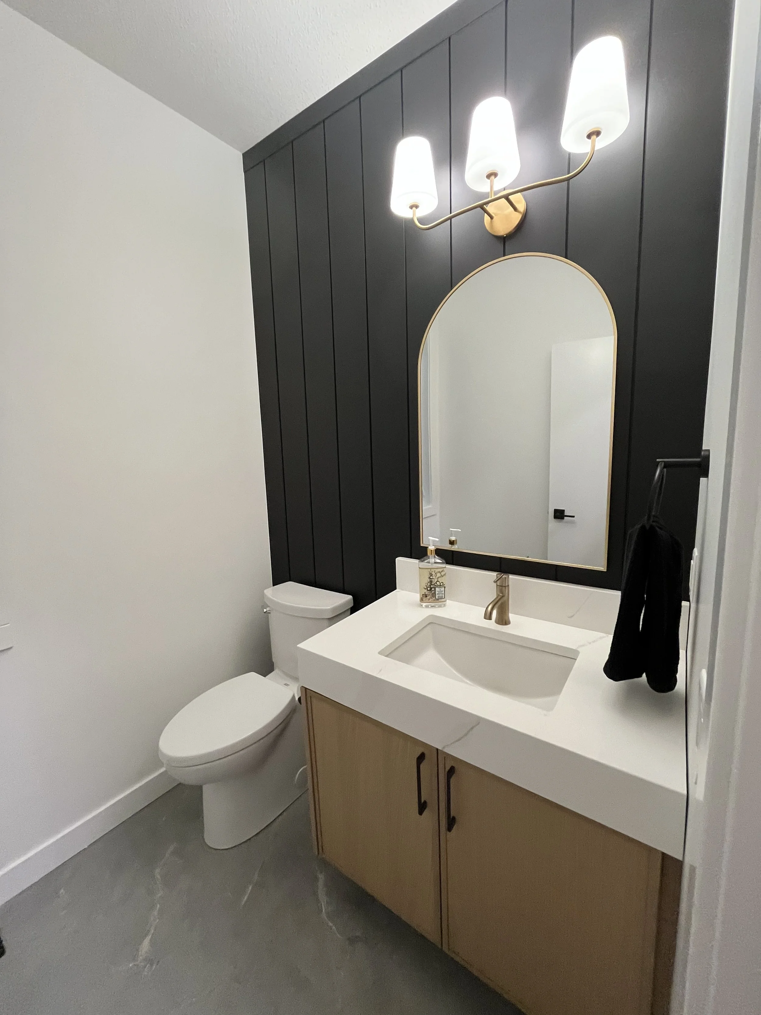 Modern bathroom with white toilet, white countertop sink, black towel, black paneled accent wall, large mirror with gold trim, and gold light fixture with three white shades.