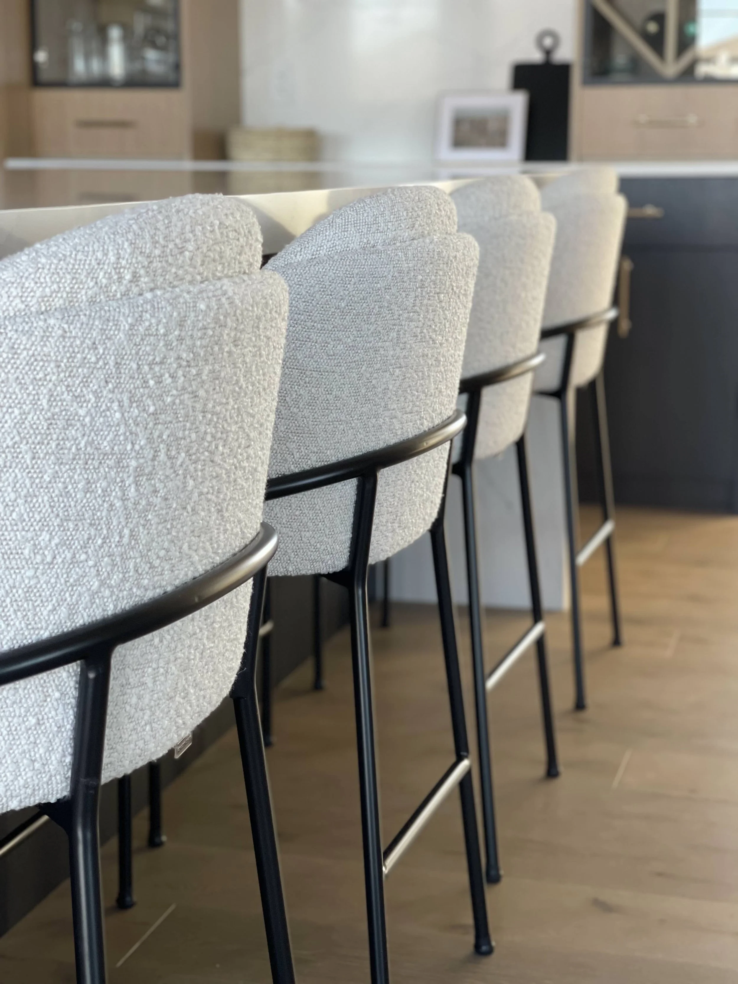 Close-up of a row of light-colored upholstered bar stools with black metal legs at a kitchen counter in a modern interior.
