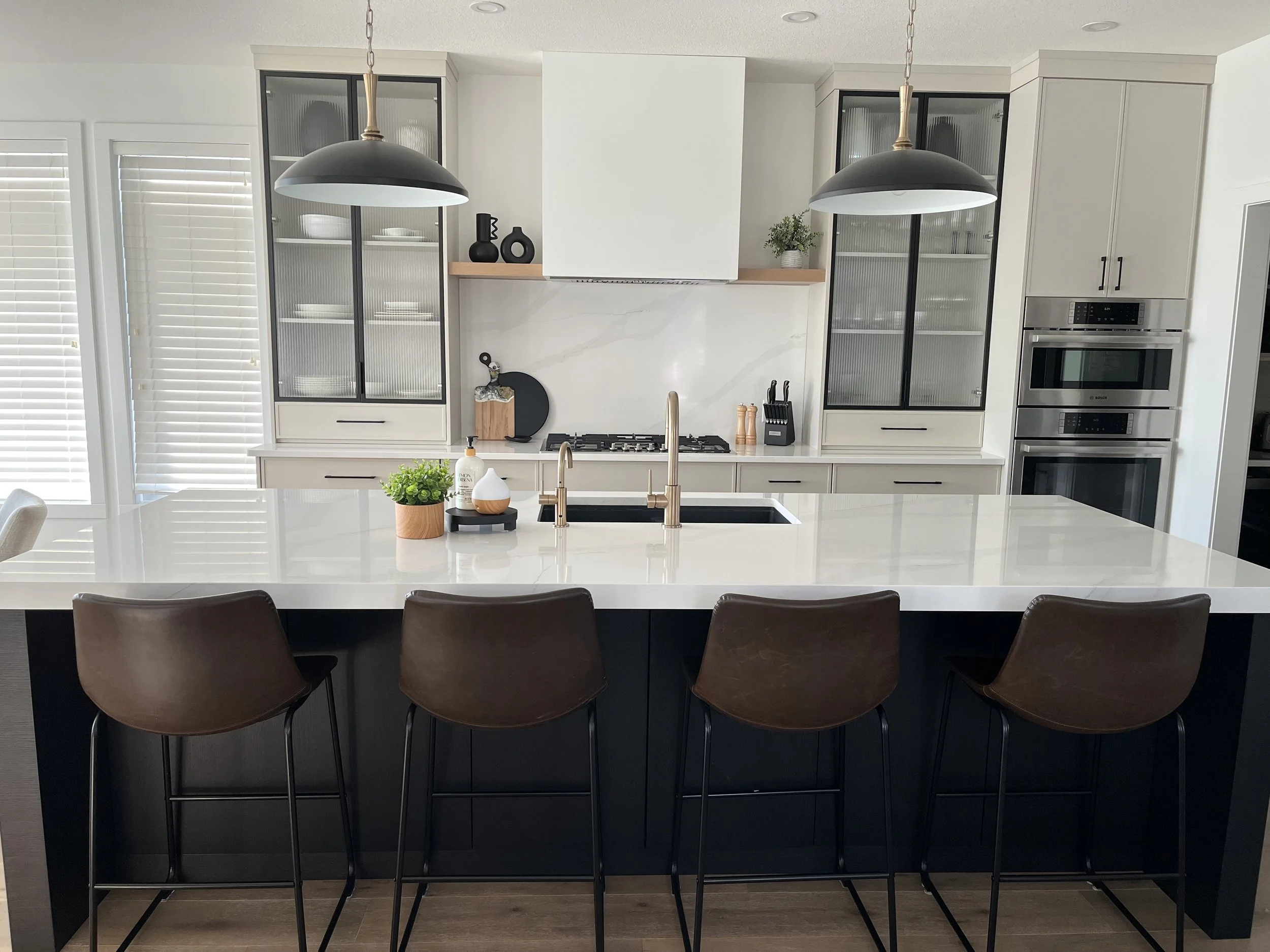 Modern kitchen with white cabinetry, a large white island with a sink and gold faucet, and black bar stools. Pendant lights hang above the island. Appliances like double ovens and a gas stove are built into the cabinetry. Decor includes plants, vases