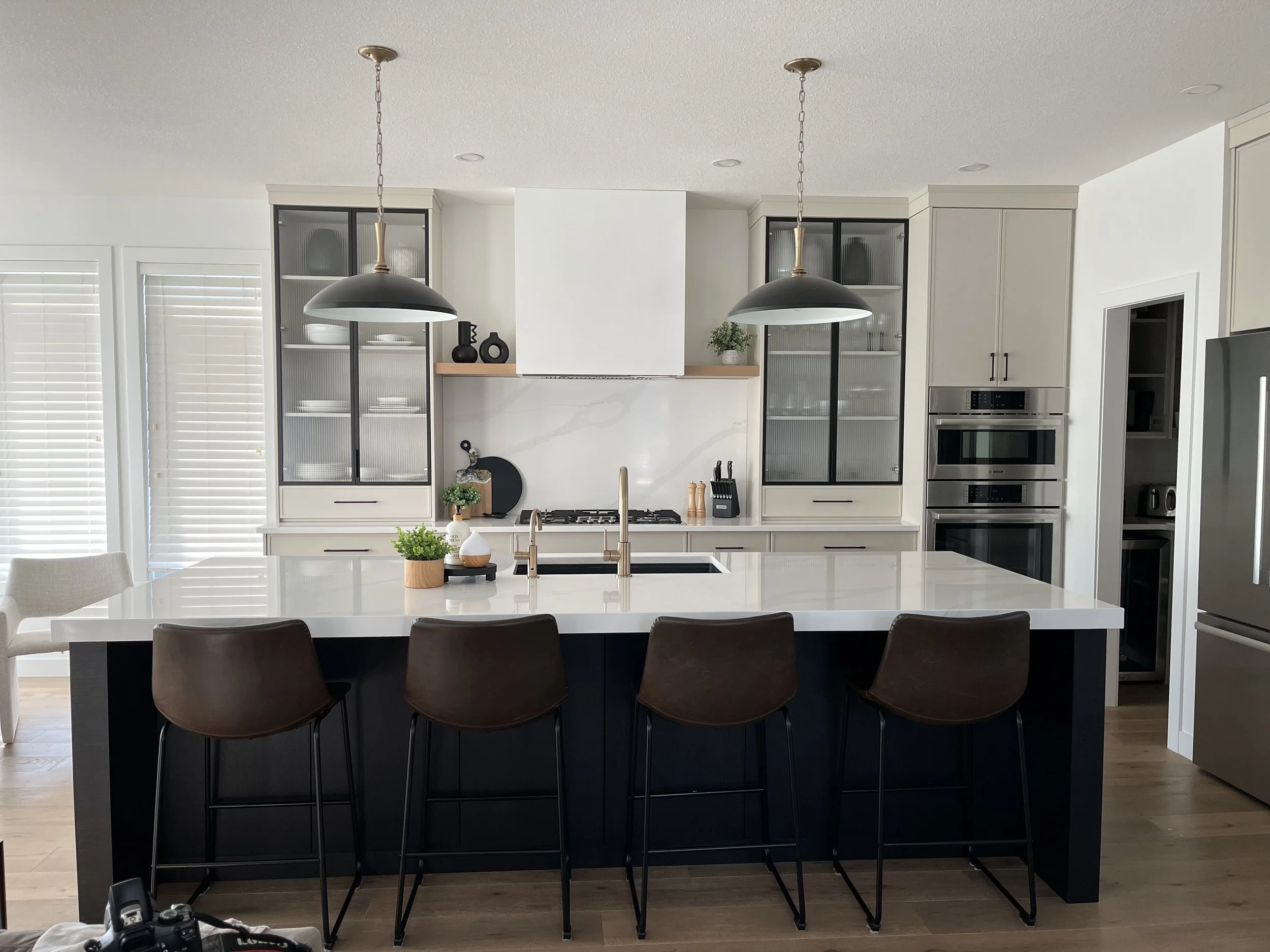 Modern kitchen with white cabinetry, black hardware, and stainless steel appliances. Large island with white quartz stone countertop and four brown chairs. Two pendant lights hang over the island. oversized pendant lights. renovation to existing home