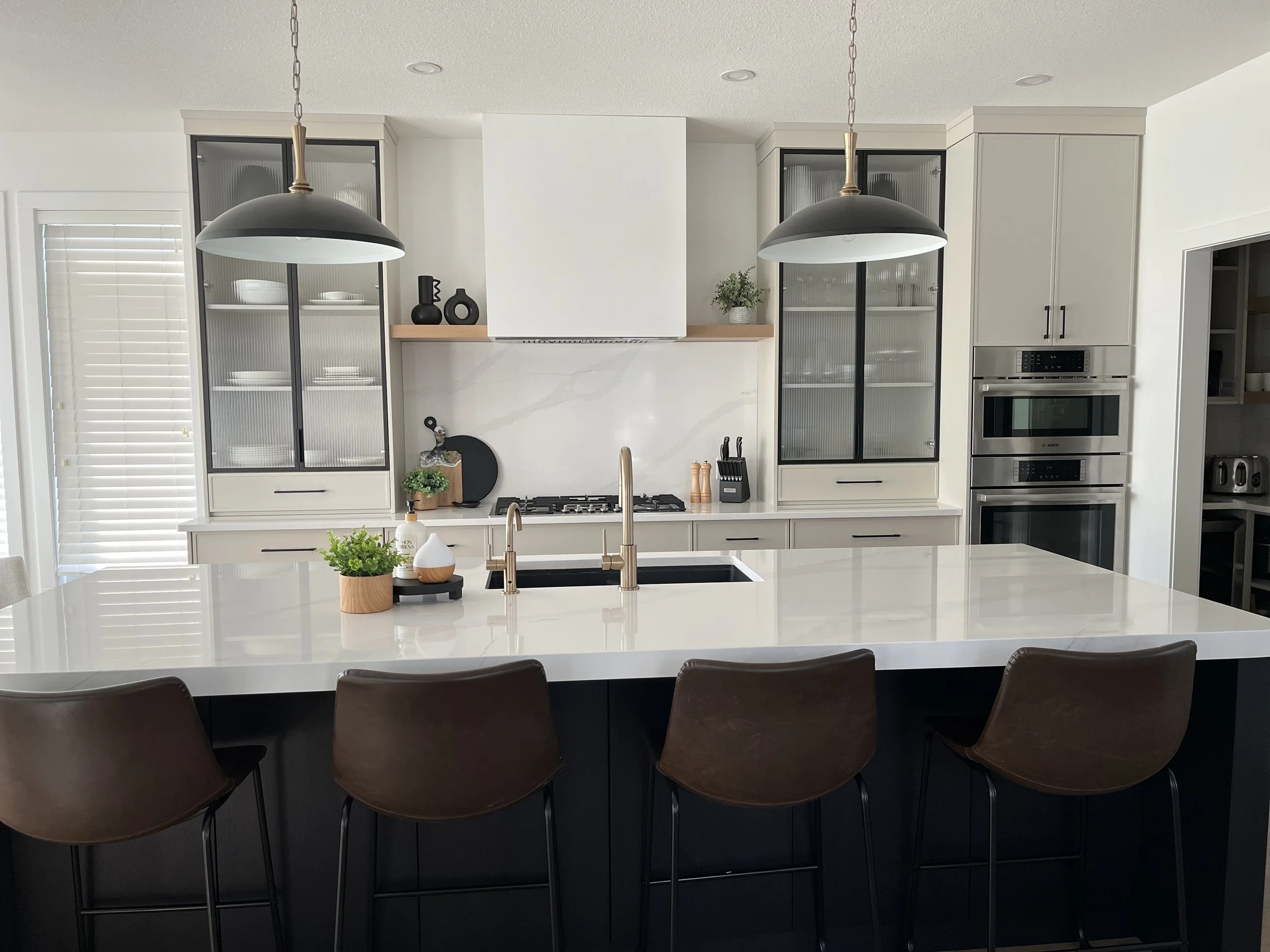 Modern kitchen with white cabinetry, black accents, built-in ovens, a large island with a white countertop, black bar stools, and black pendant lights.