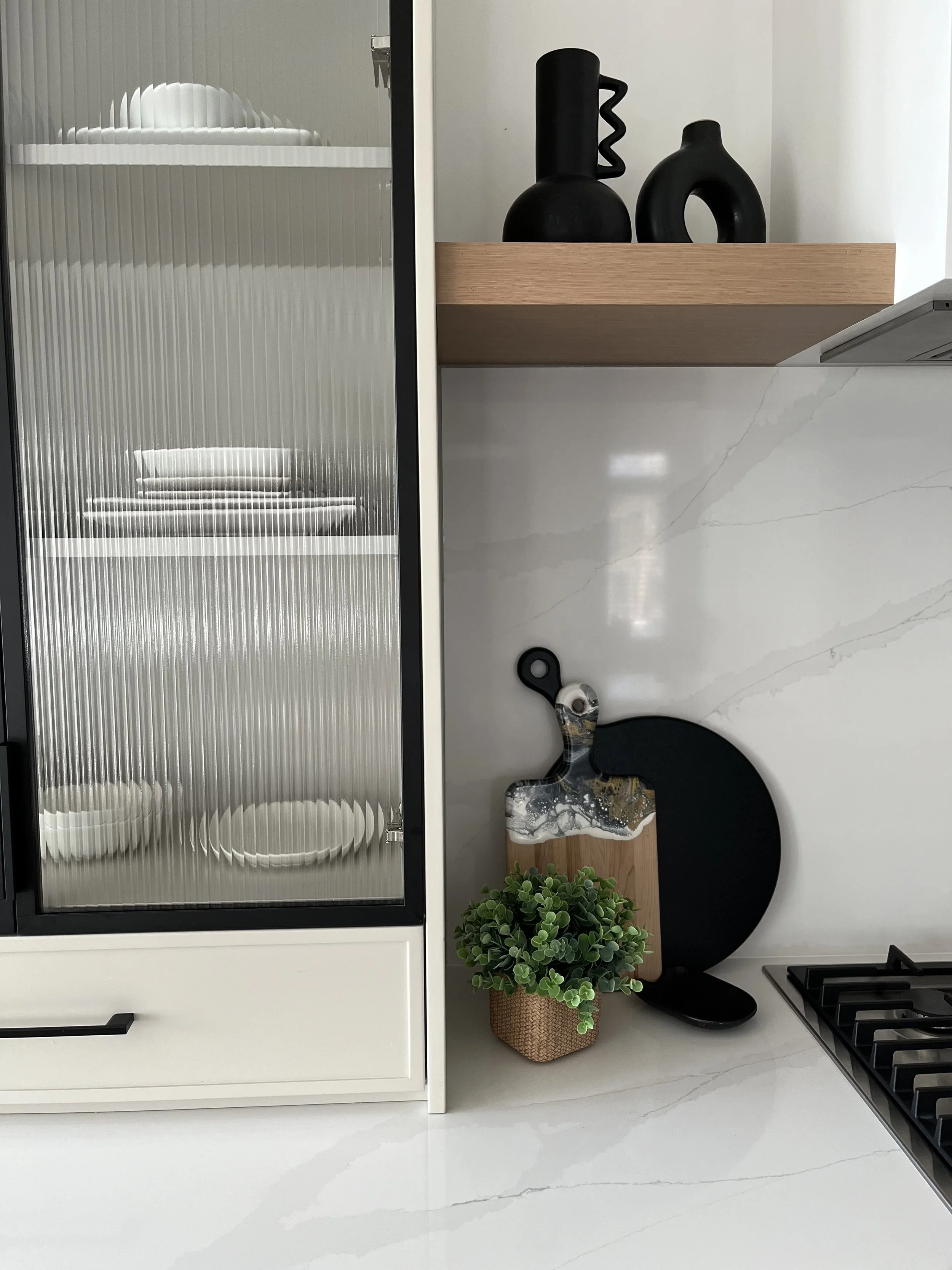 Modern kitchen with a white marble countertop, black decorative vases on a wooden shelf, a cutting board with a marbled handle, a small potted plant, and a black round chopping board.
