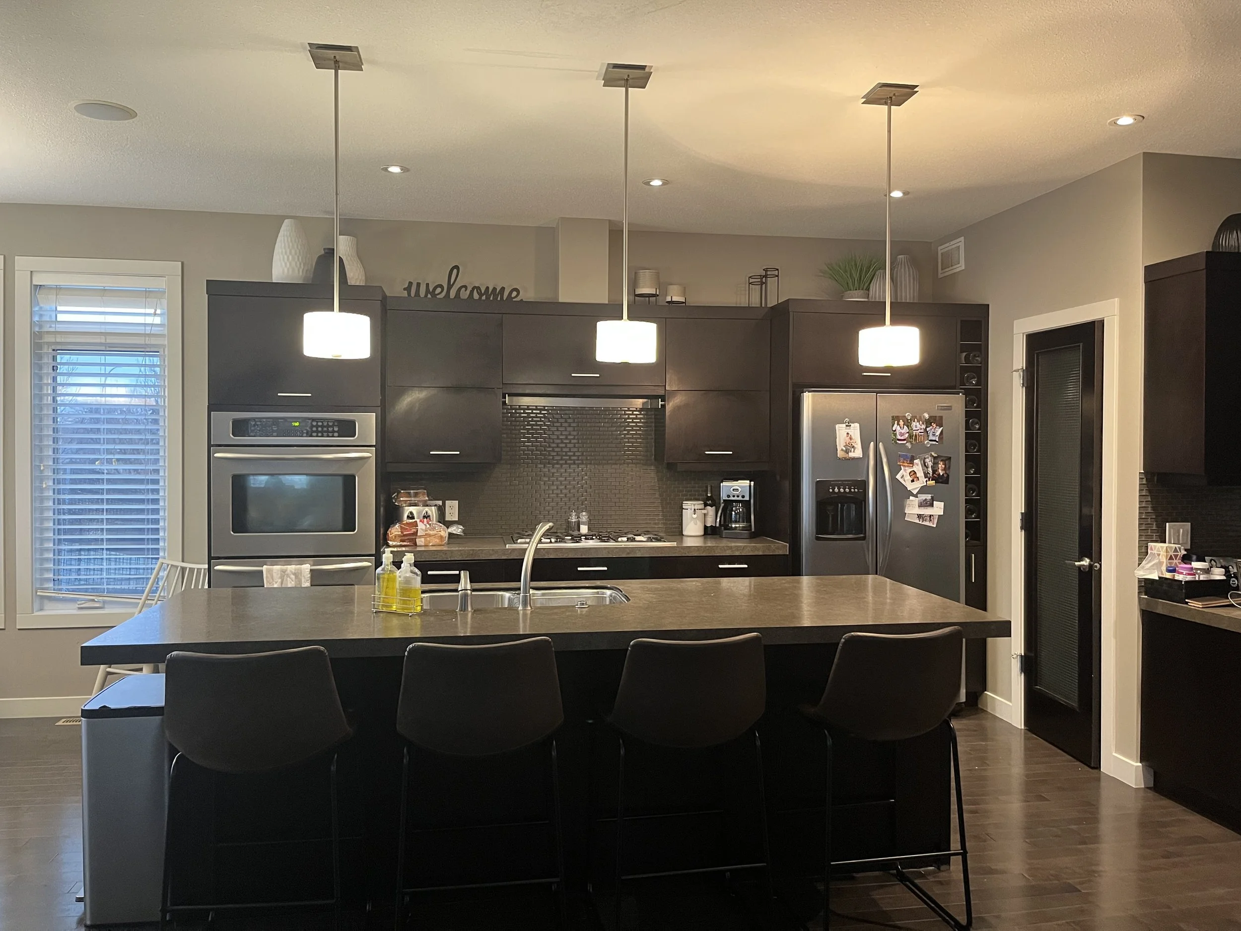 Modern kitchen with dark cabinetry, stainless steel appliances, and a large island with four black chairs. Pendant lights hang above the island. There are windows with white blinds on the left and various small decorative items on top of the cabinets