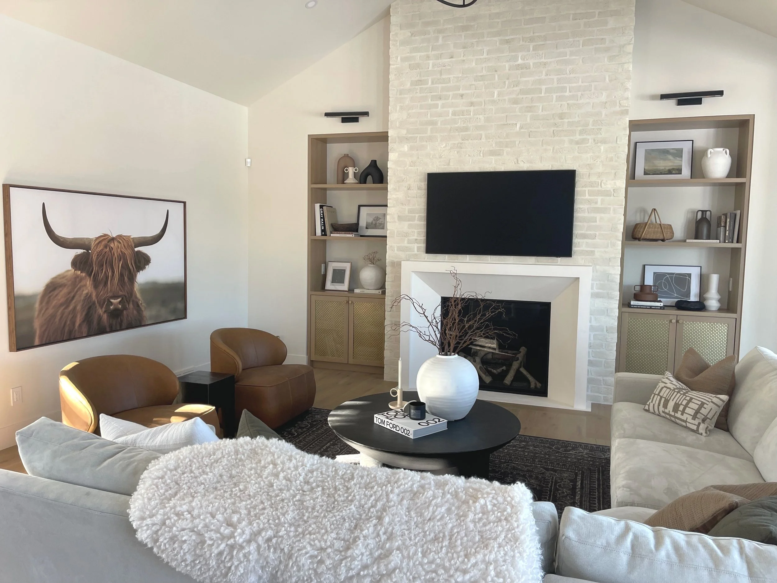 Living room with white sofa, round black coffee table, brown chairs, built-in shelves, and a white brick fireplace with a mounted flat-screen TV. Decorative items include a large white vase with twigs, framed art, books, and vases. Large framed photo of a Highland cow with horns on the left wall.