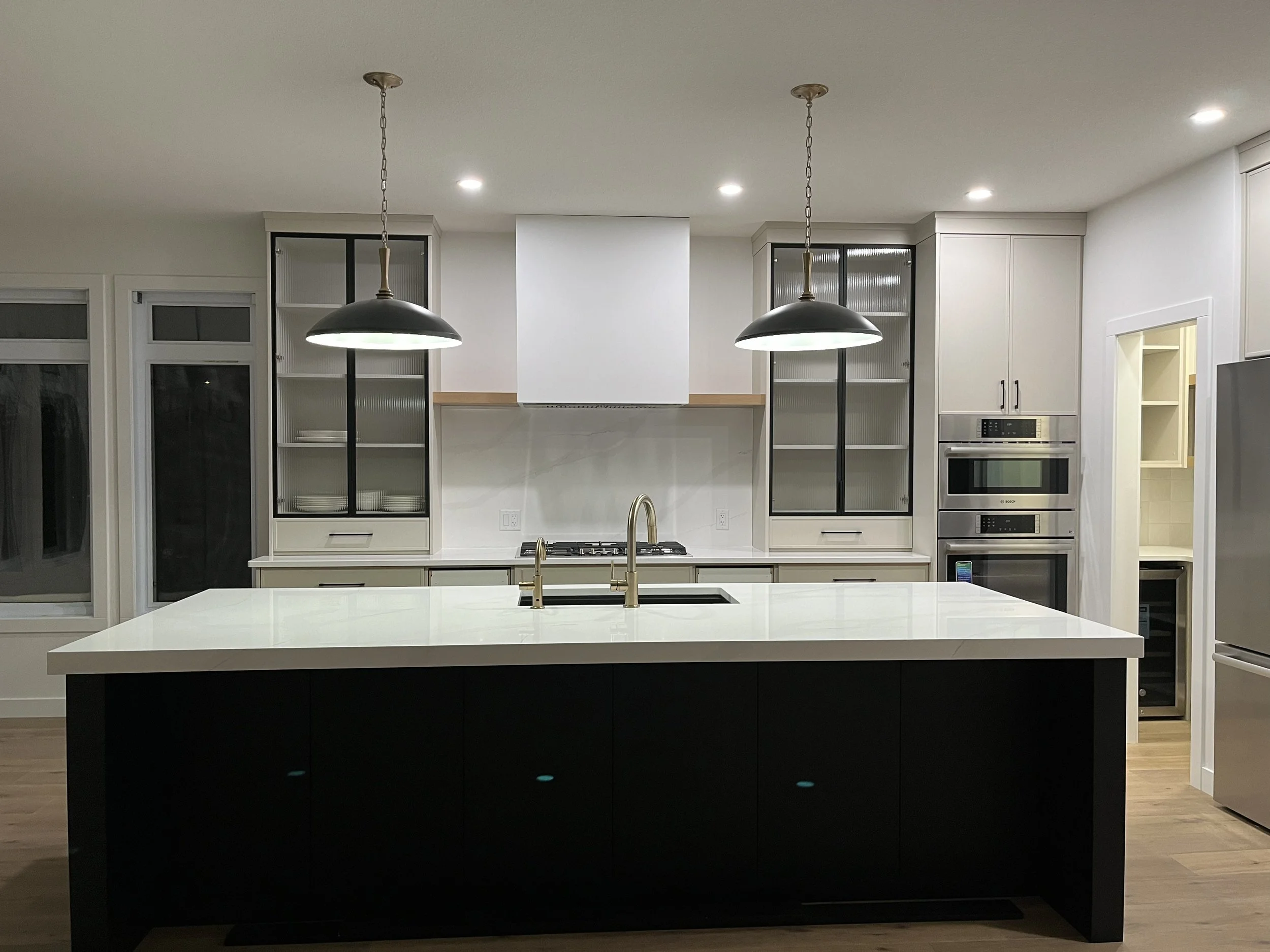 Modern kitchen with white countertops, black island, pendant lights, built-in ovens, stainless steel refrigerator, and cabinets with glass doors.