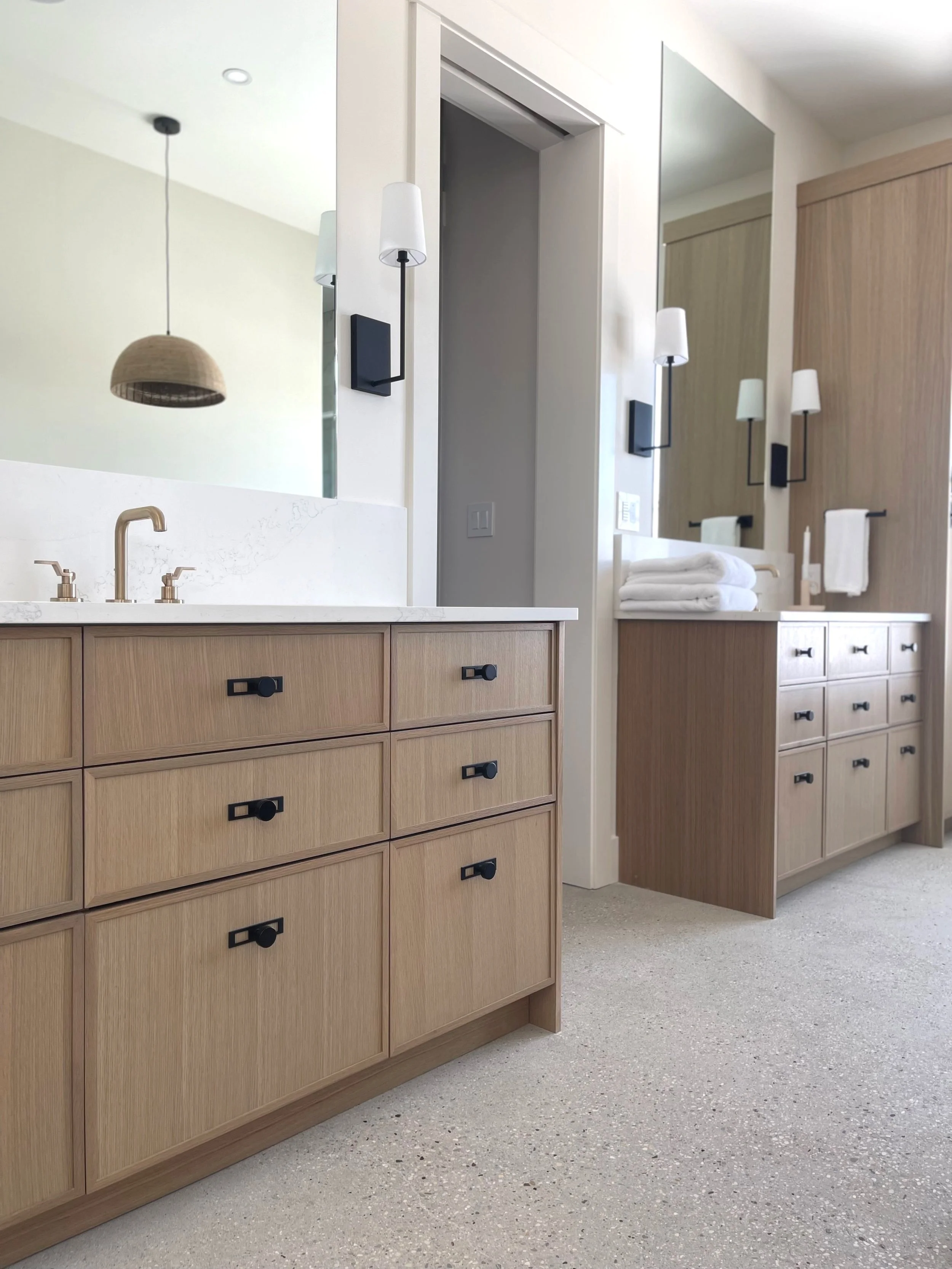 Modern bathroom with light wood cabinets, white countertop, black hardware, large mirror, wall-mounted light fixtures, folded white towels, and beige floor.