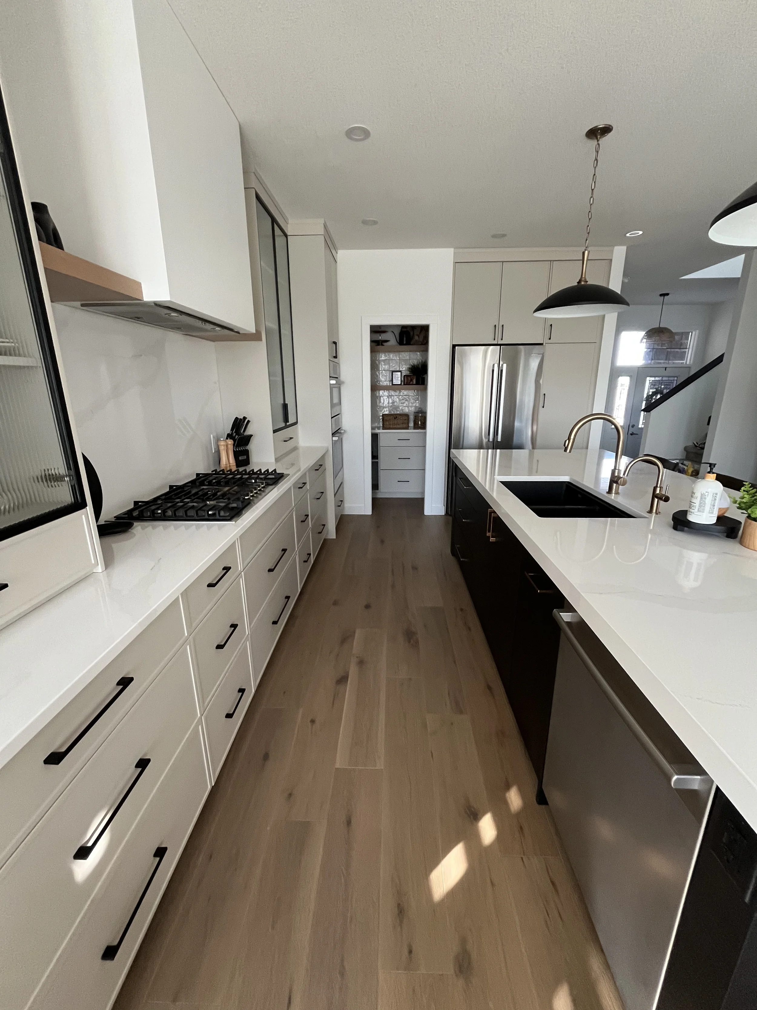 Modern kitchen with white cabinets, black hardware, a large island with a sink and gold fixtures, stainless steel appliances, and wooden flooring.
