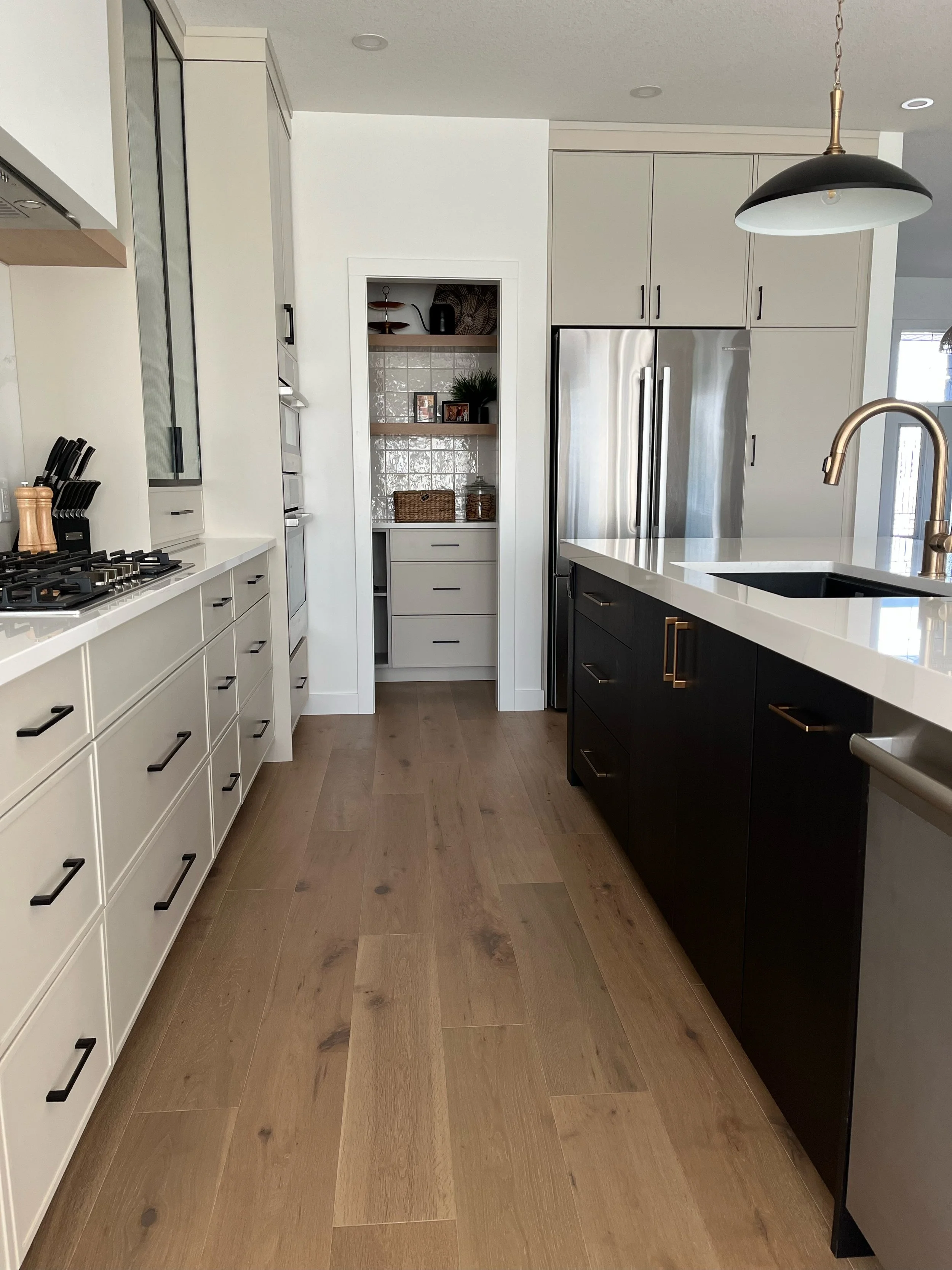 Modern kitchen with light-colored cabinets, black hardware, a stainless steel refrigerator, a black island with a brass handle, and a wooden floor. There is a small built-in open shelf area with decorations and framed photos.