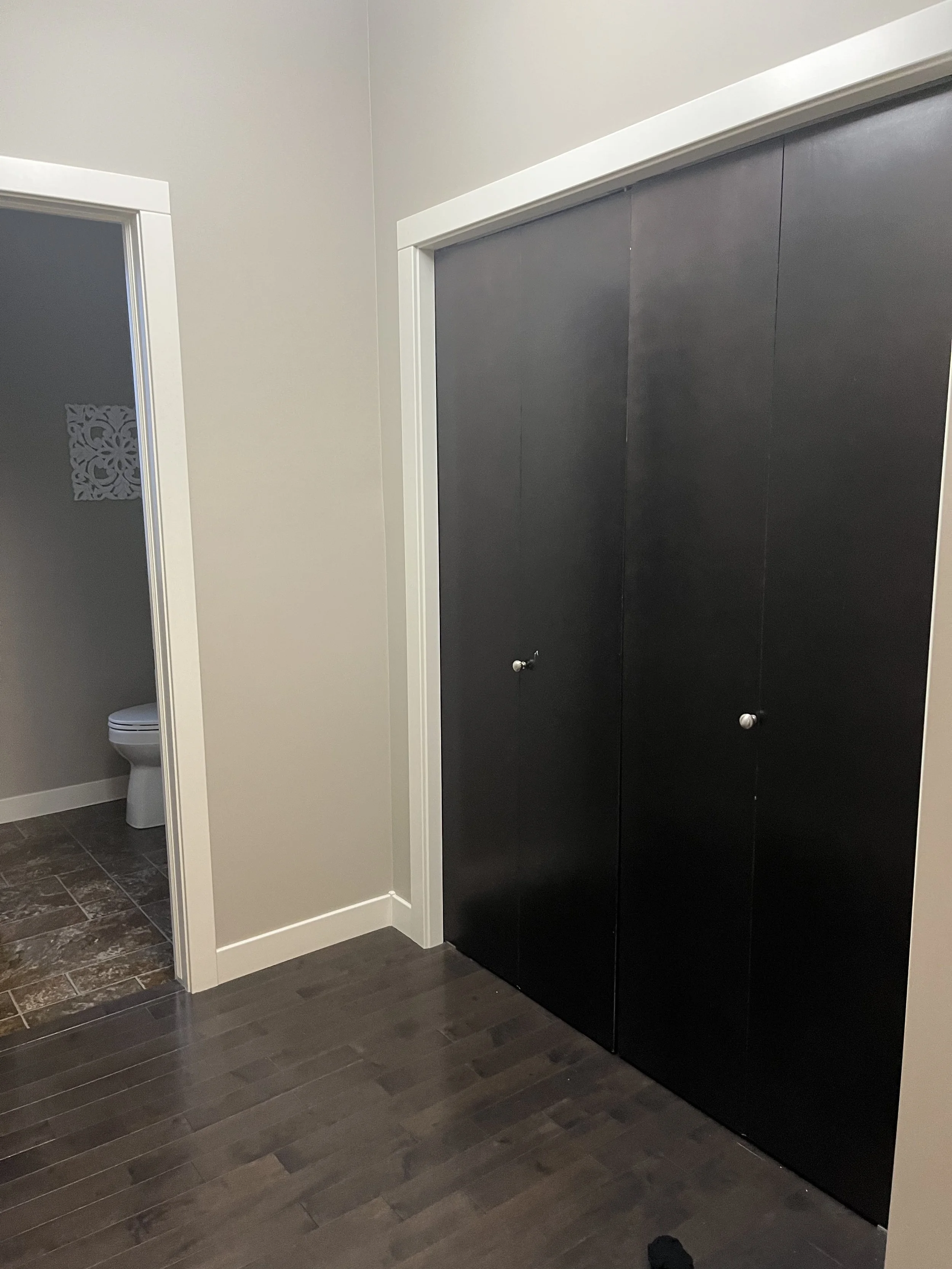 Interior view of a room with dark wooden floor, beige walls, and a closed black closet door with knob. In the background, a partially visible bathroom with white toilet and decorative wall features.