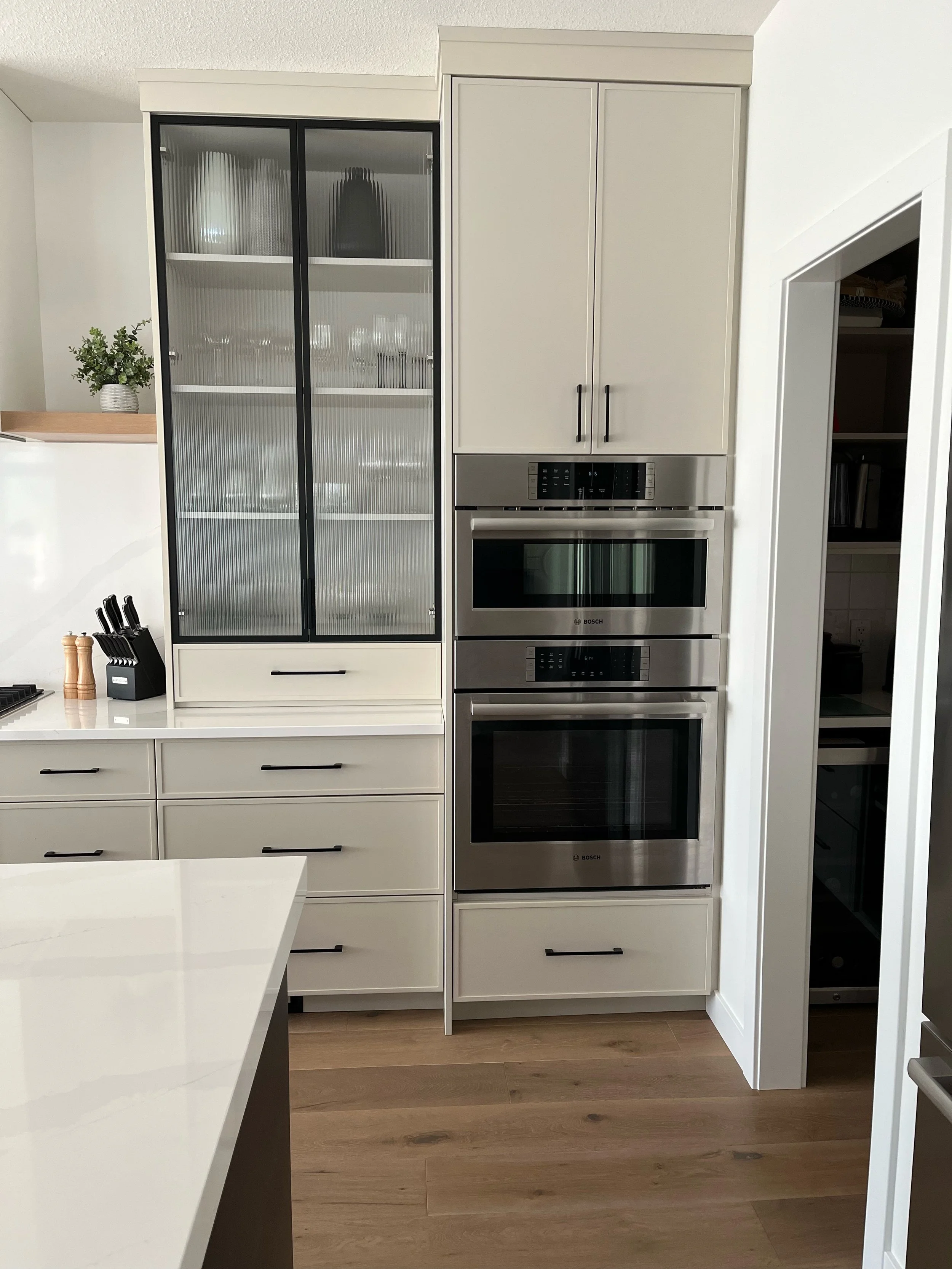 Modern kitchen with white cabinetry, stainless steel built-in oven and microwave, glass-front cabinet, and wooden flooring.