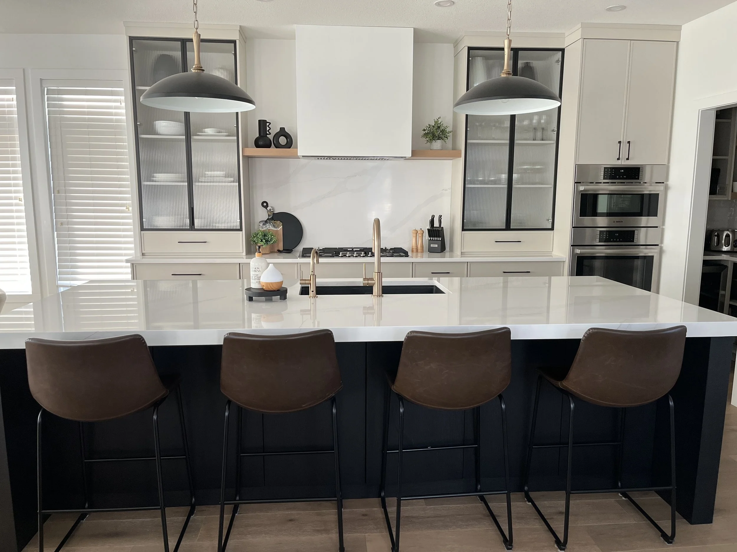 Modern kitchen with black island, white countertops, and brown bar stools. Features built-in oven, gas stove, glass-front cabinets, pendant lights, and minimal decor.