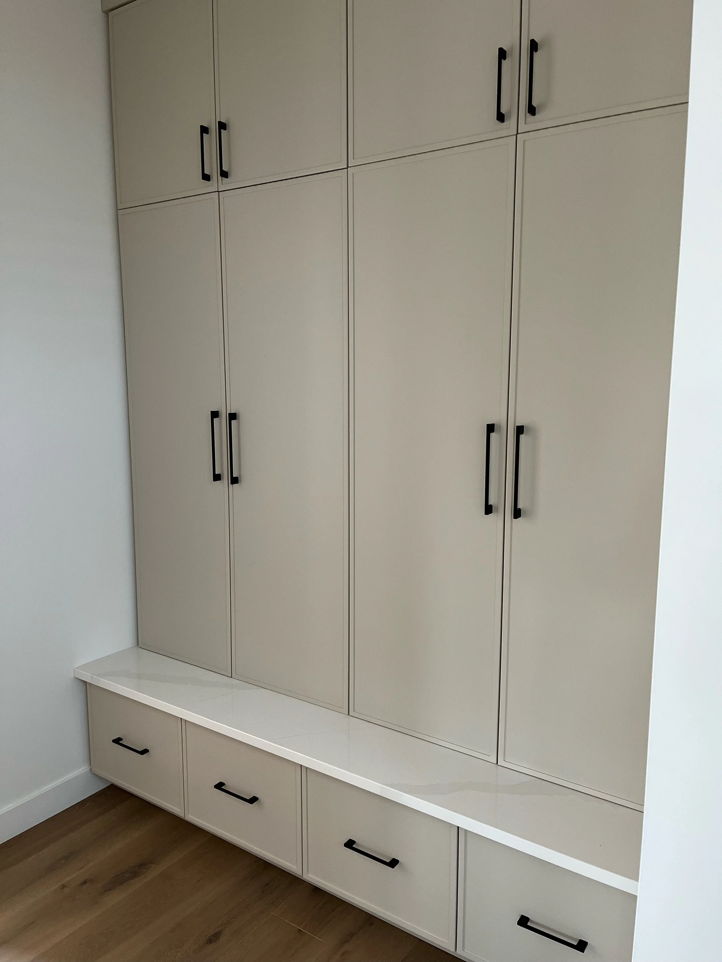 White built-in storage cabinet with black handles, upper and lower sections, located in a corner with hardwood floor.