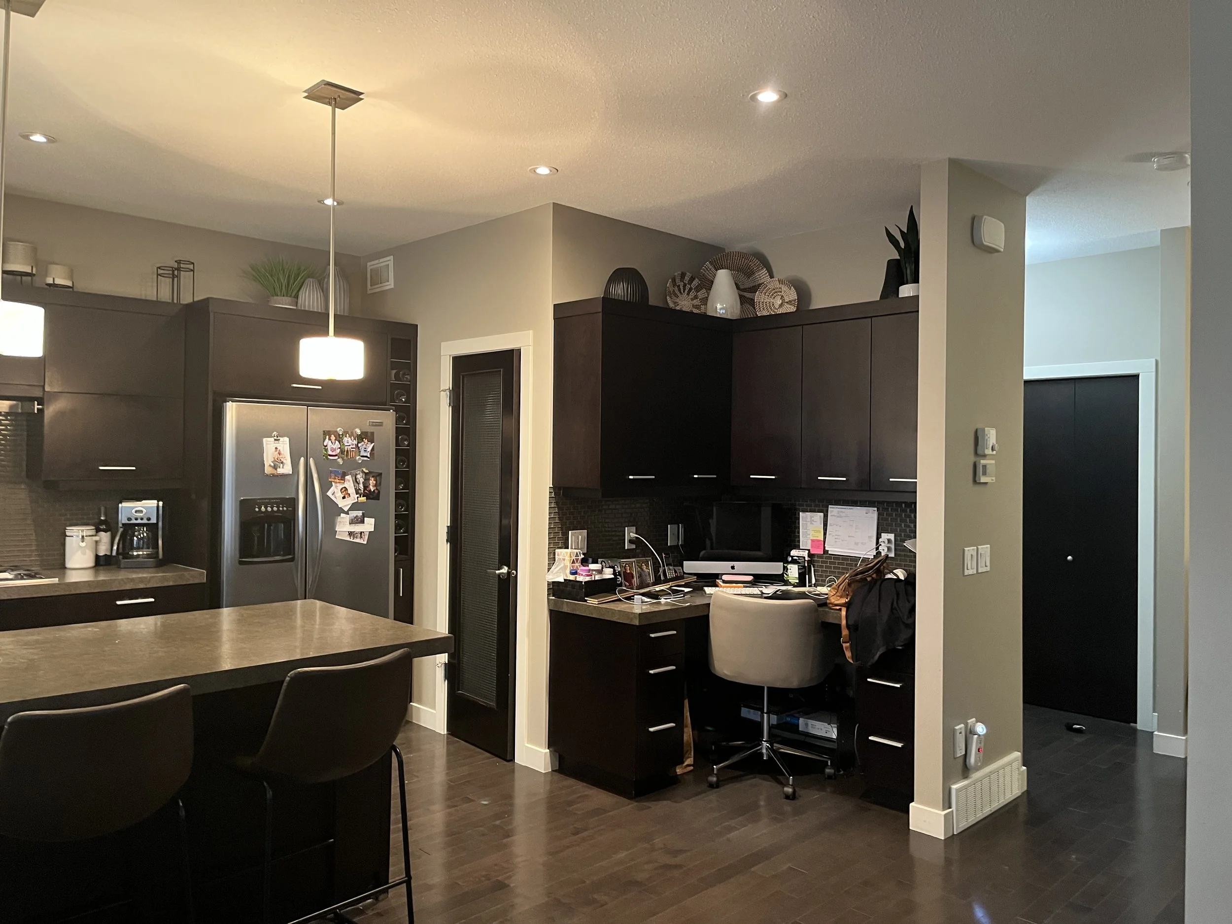 Modern kitchen with dark wood cabinetry, stainless steel refrigerator, and a small home office desk with a beige swivel chair.