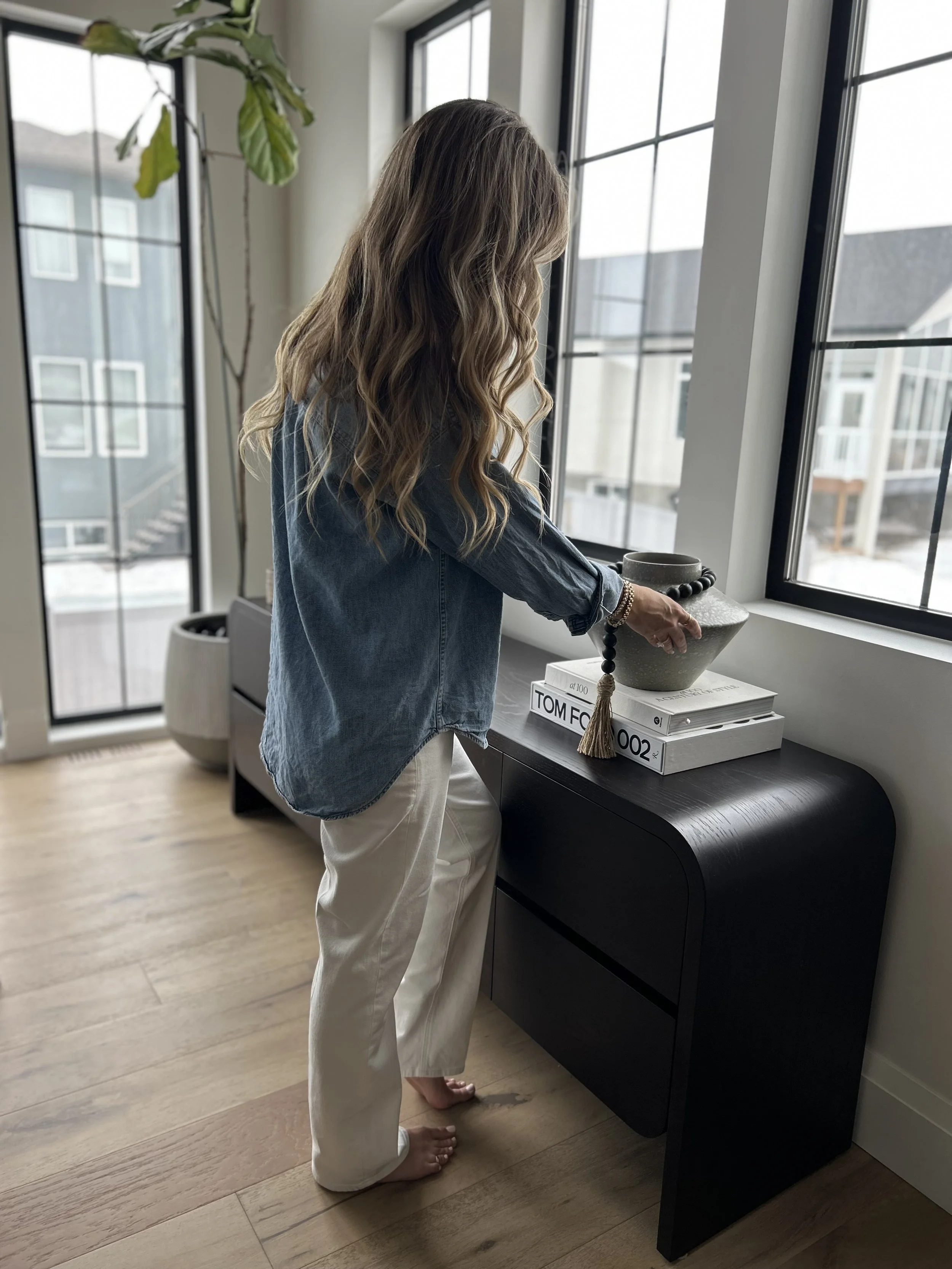 Woman with wavy hair in a denim shirt and white pants arranging decorative vases and books on a black side table near large windows in a modern apartment.