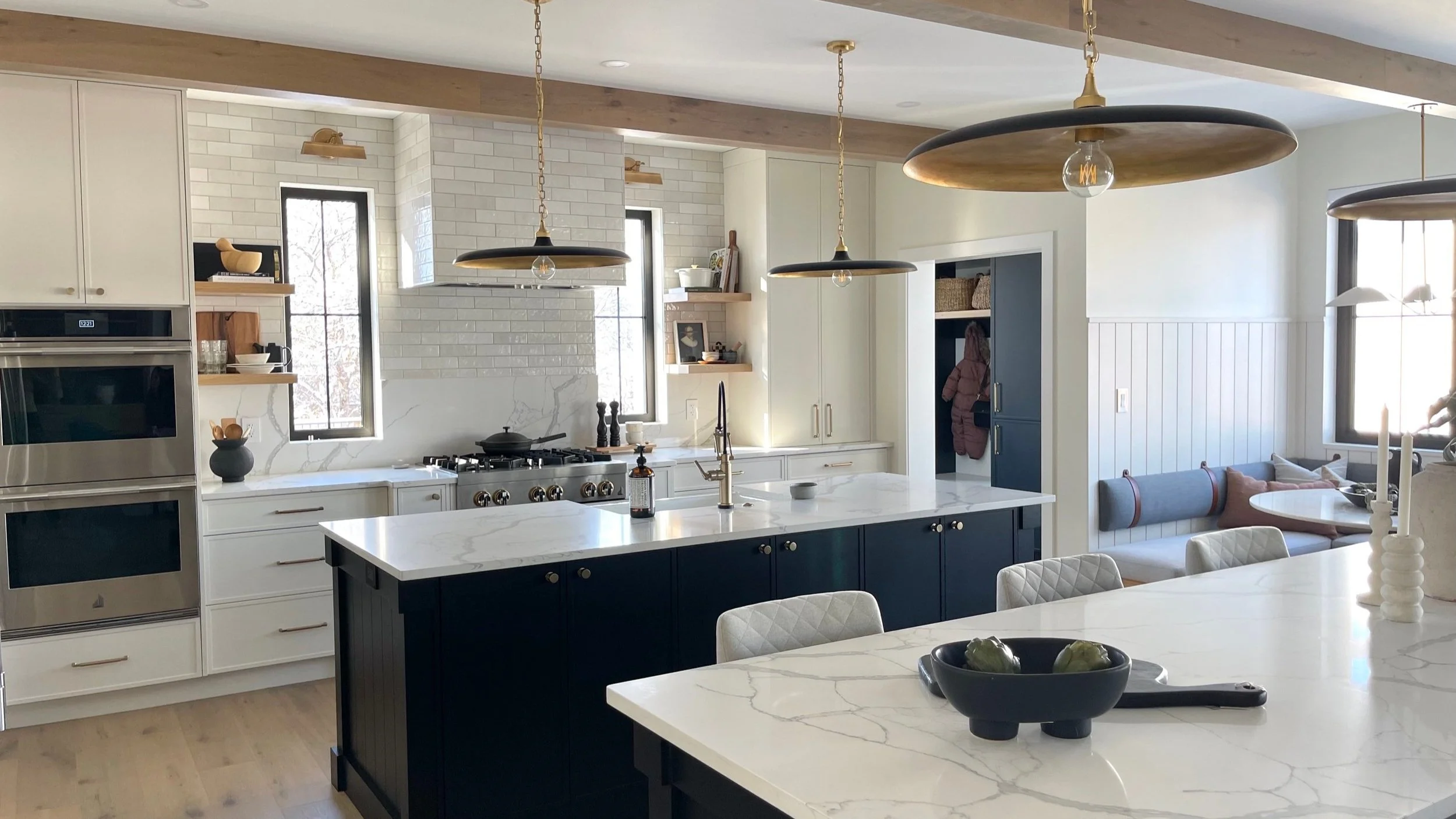 Modern kitchen with white cabinetry, navy blue island, white marble countertops, and gold pendant lighting, with a seating nook and natural light through windows.