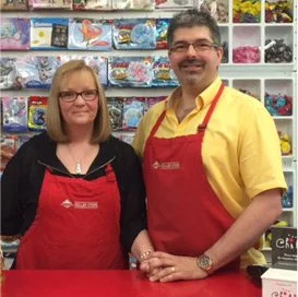 A woman and a man standing at a store counter wearing red aprons, smiling. The background shows shelves with various colorful items.