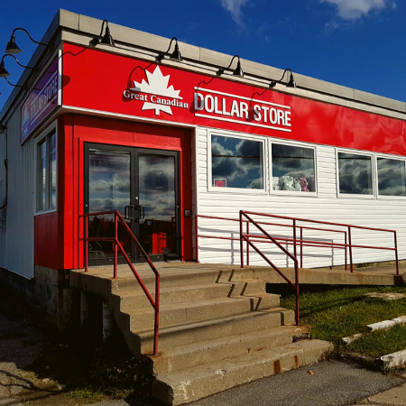 Exterior of a Dollar Store with red and white siding, a red and white sign reading 'Great Canadian DOLLAR STORE', glass windows, a main entrance door, and concrete stairs with red railings.