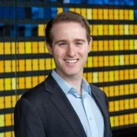 A man in a suit smiling in front of a background of yellow and blue server racks or data storage units.