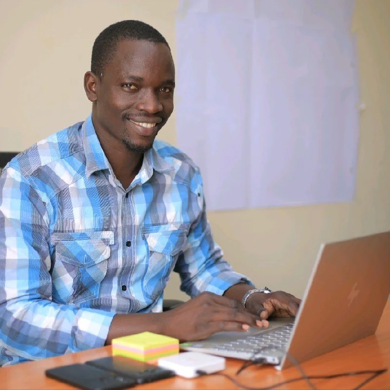 Smiling man in a checkered blue shirt working on a laptop at a desk with sticky notes and a smartphone.
