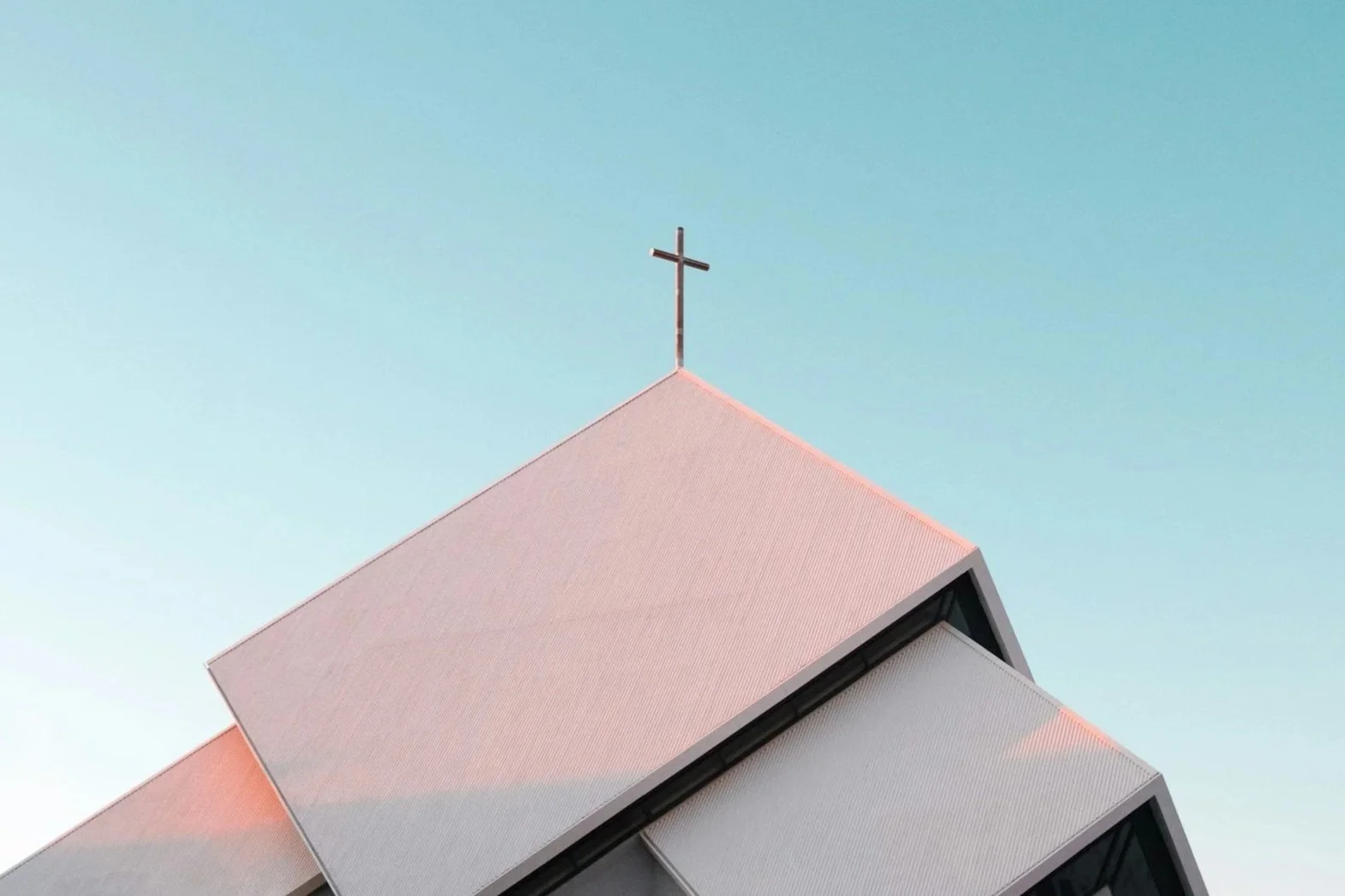 Close-up of church roof with a cross at top against a pale blue sky.