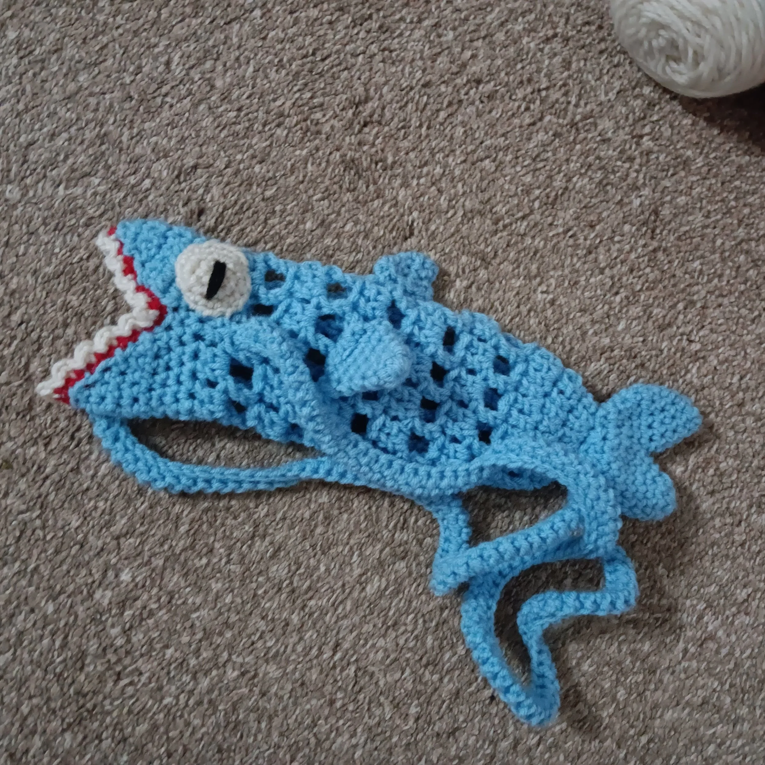 Handmade crocheted blue shark-shaped bag with an open mouth, white eye with black pupil, and a small dorsal fin, lying on a beige carpet.