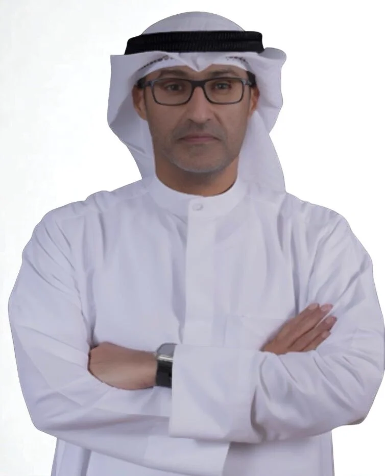 A man wearing traditional Middle Eastern attire, including a white kandura, a white headscarf (ghutra), and a black agal, with glasses and a neutral expression.